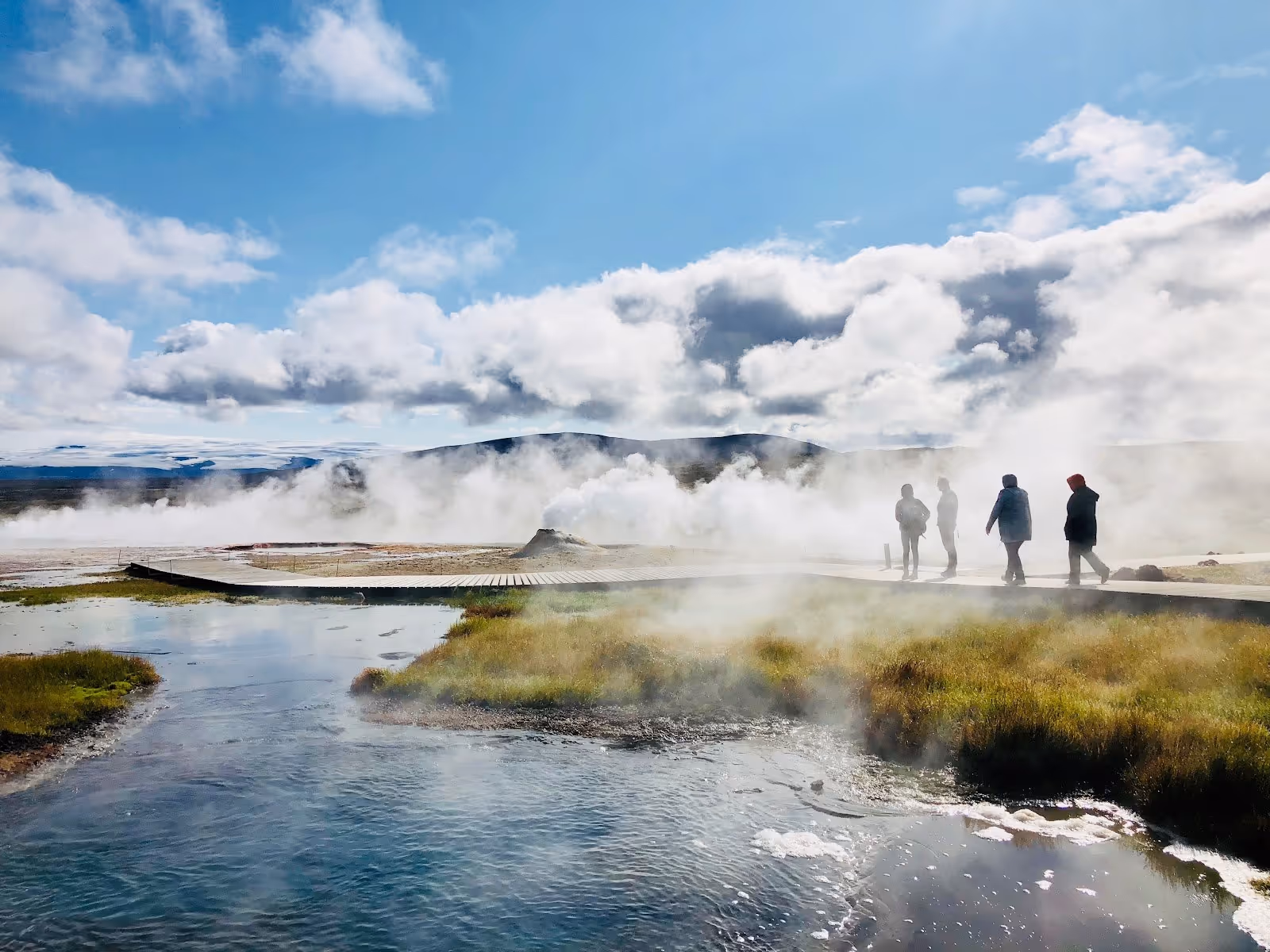 Wooden boardwalk crossing a steaming geothermal hot spring area with hot water pools and hills under a partly cloudy sky at Hveravellir geo.