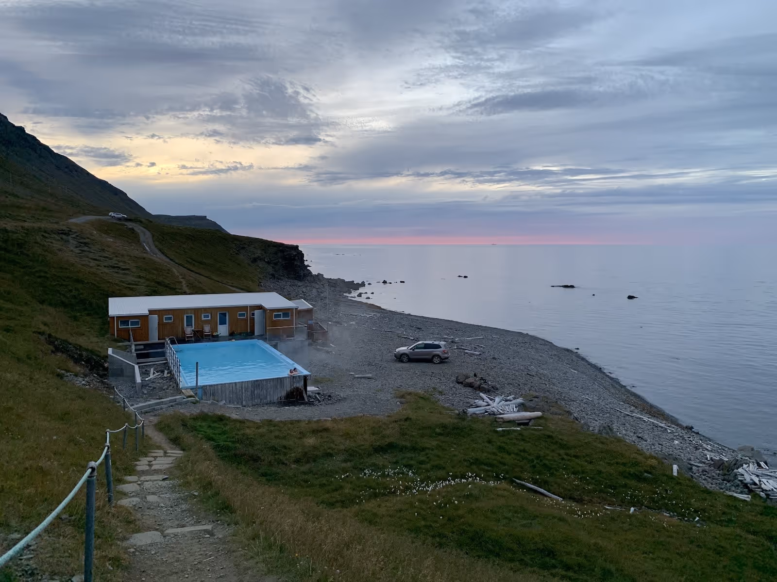 Aerial view of Krossnes Swimming Pool near Nordurfjordur, Iceland, showing a rectangular pool beside the rocky coastline with steam rising.