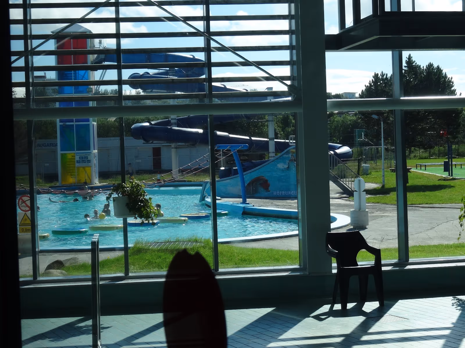 View through large windows to outdoor water slide and hot spring pool with people playing and greenery around in Reykjavik Iceland.
