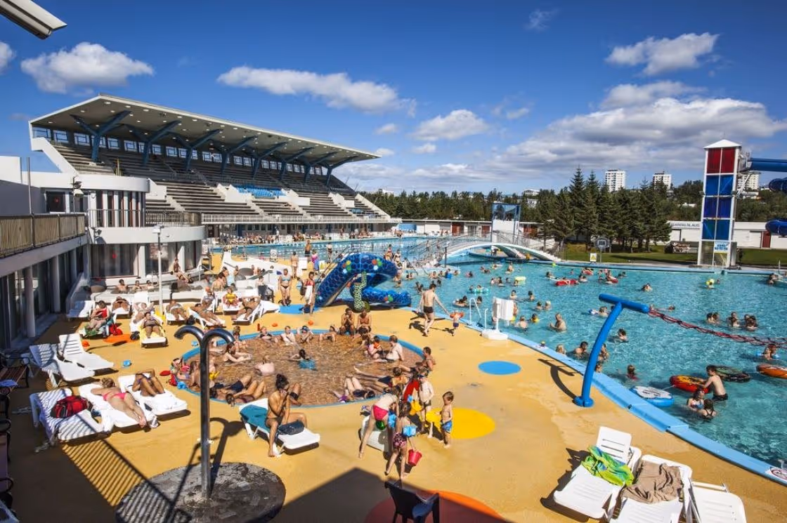 Outdoor hot spring pool at Laugardalslaug in Reykjavik Iceland with many people swimming and lounging on chairs under a blue sky.