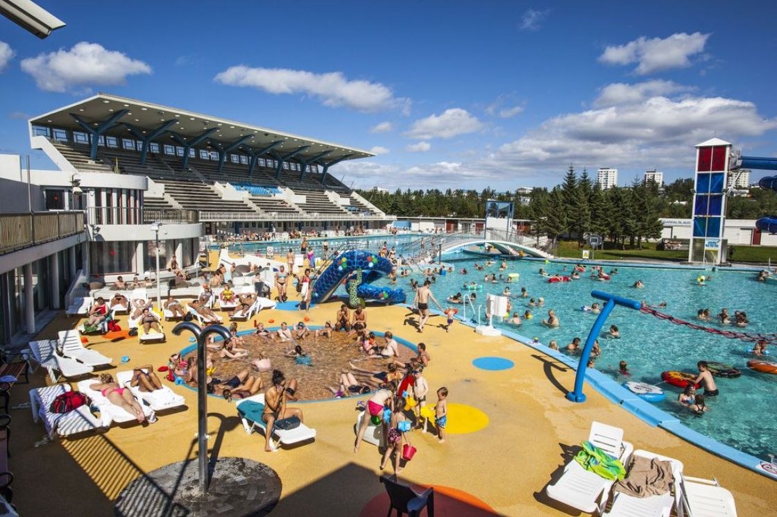 Outdoor hot spring pool at Laugardalslaug in Reykjavik Iceland with many people swimming and lounging on chairs under a blue sky.
