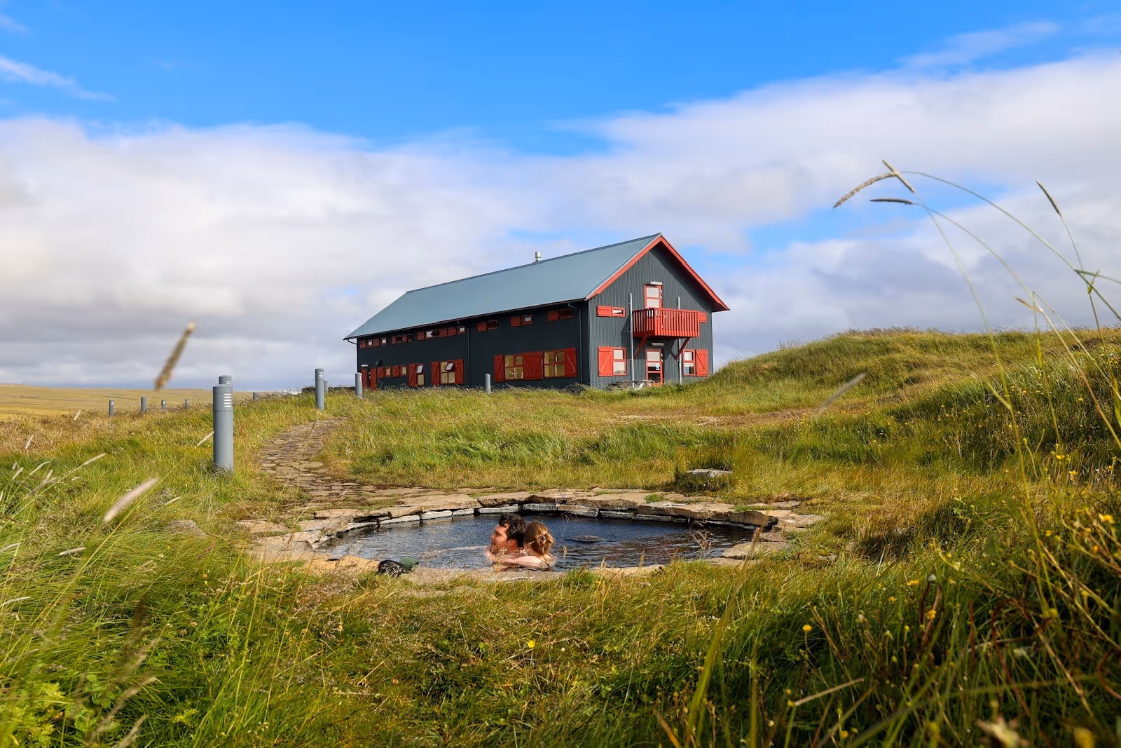 Two people relaxing in a small stone-bordered hot spring pool at Laugarfell Hot Springs near Egilsstadir Iceland under a cloudy blue sky.