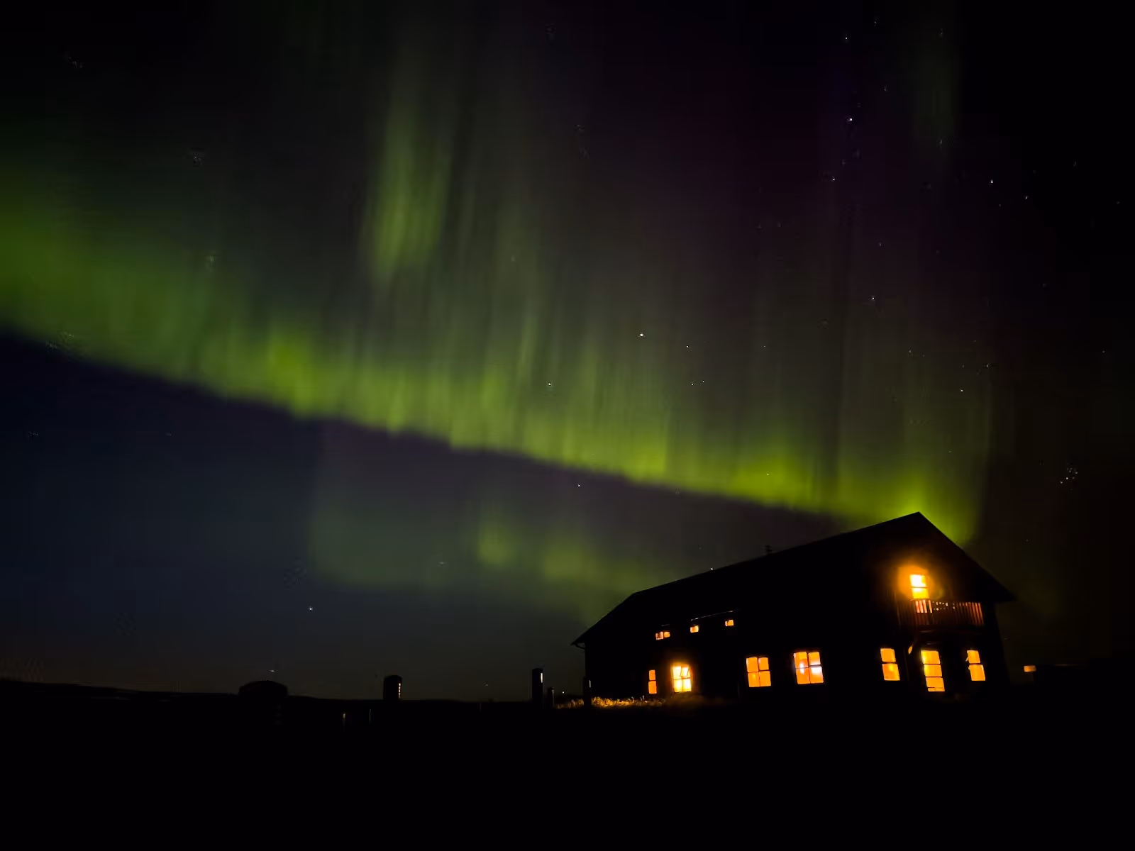Dark silhouette of a building lit from inside with northern lights glowing bright green and purple in the night sky above in Iceland.