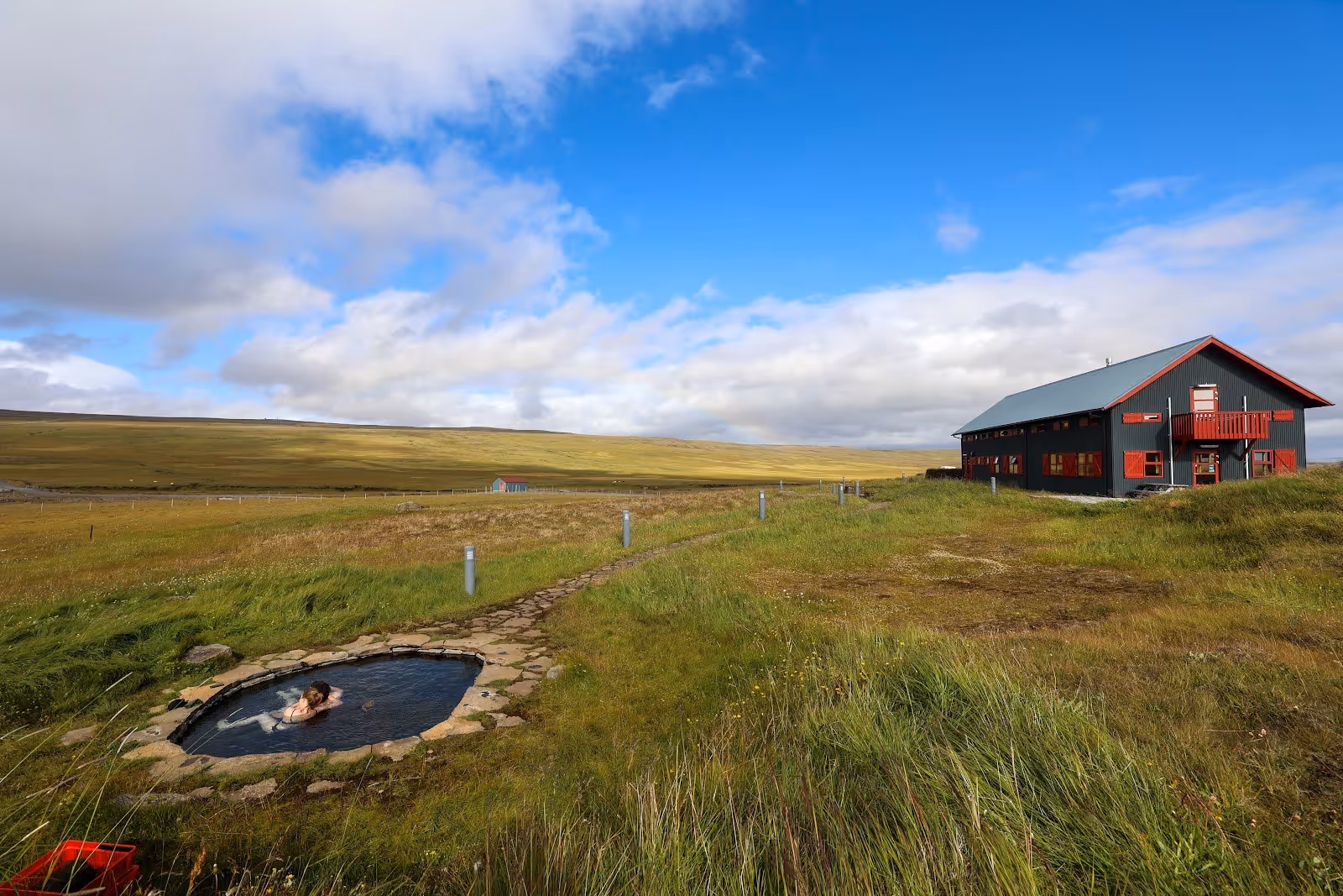 Single person enjoying the outdoor hot spring pool at Laugarfell Hot Springs with a wide grassy landscape and partly cloudy sky in Iceland.