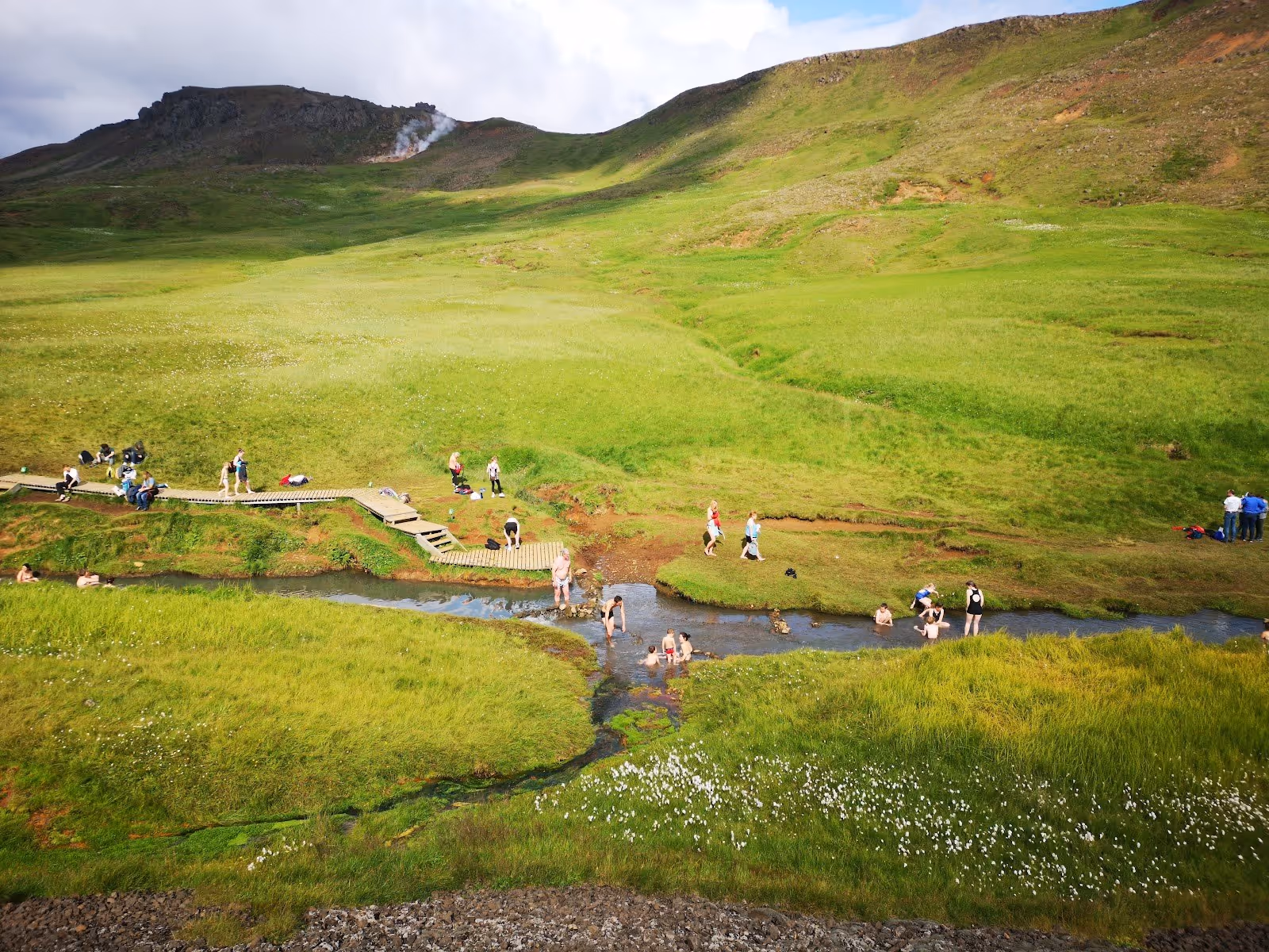 Groups of people enjoying soaking in shallow natural hot spring pools surrounded by lush green hills at Reykjadalur Thermal River in Icelan.