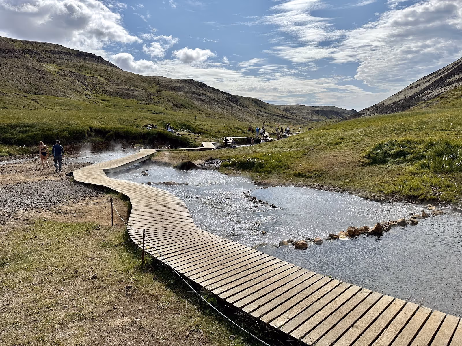 A winding wooden boardwalk beside a steaming hot spring river with visitors walking and soaking near Hverageroi in Iceland.