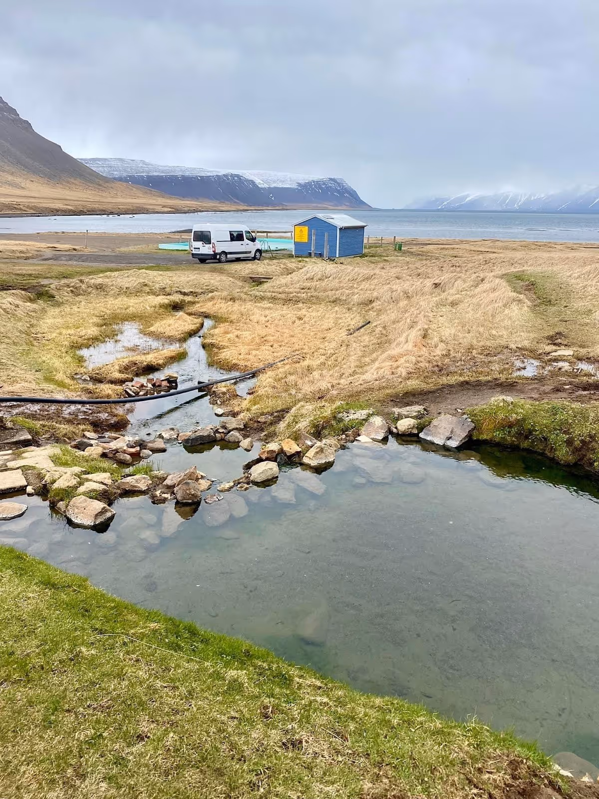 Natural hot spring pool next to small buildings, with ocean and mountain views in the background near the Reykjafjardarlaug Hot Pool in Ice.