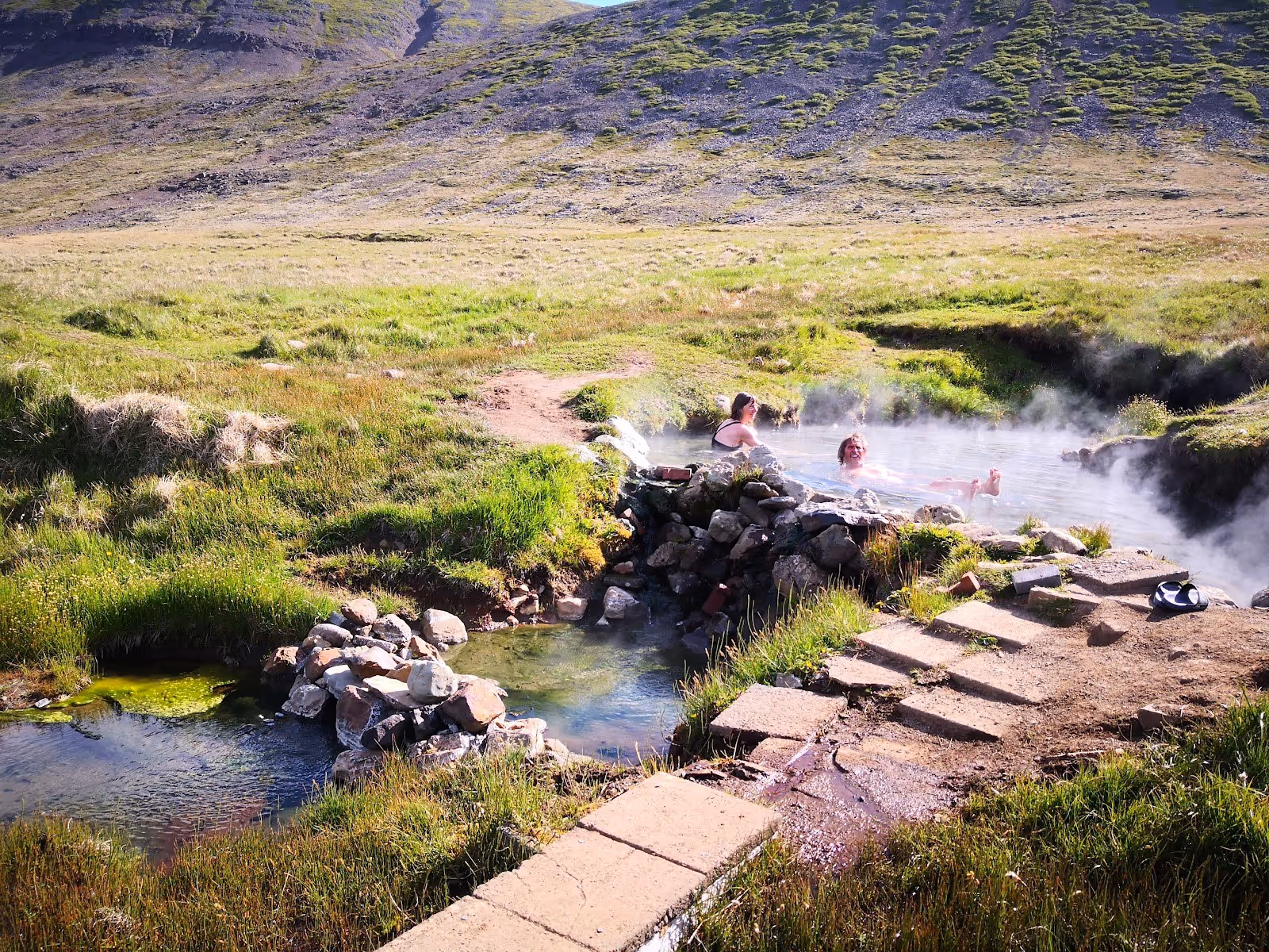 Reykjafjardarlaug Hot Pool with two people soaking in steam rising from a natural stone pool surrounded by grassy terrain.