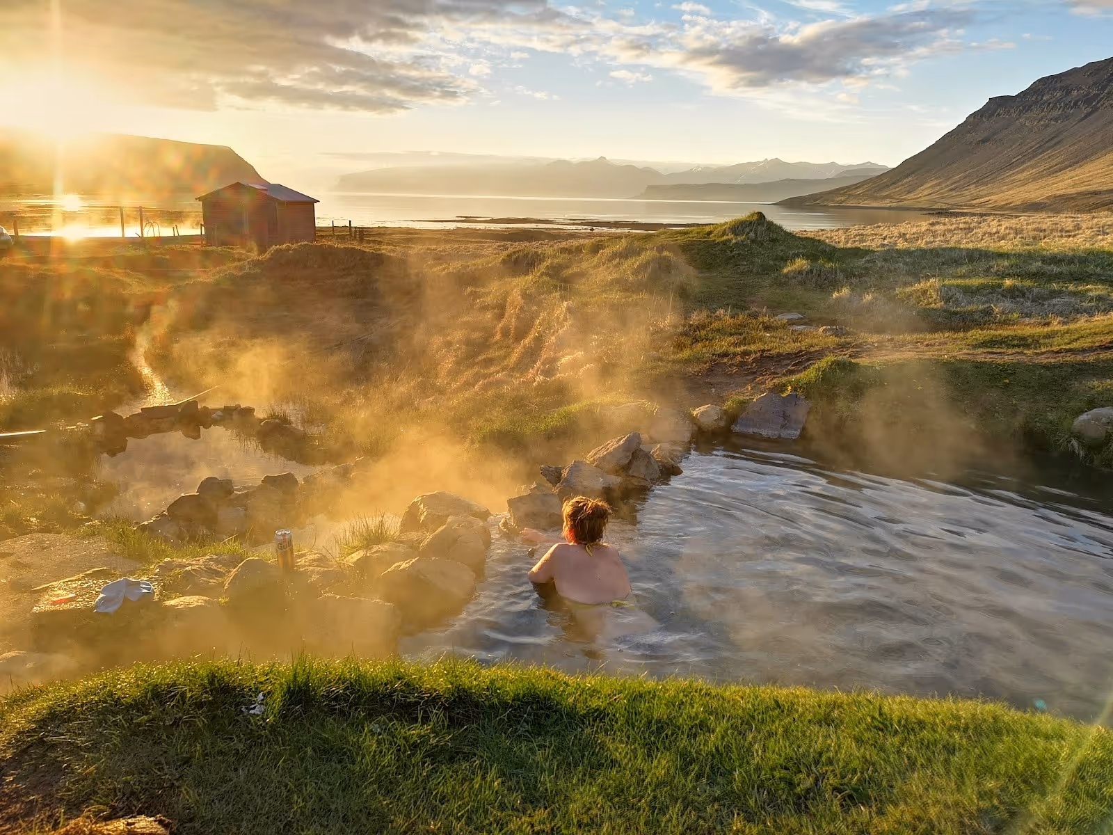 Sunset light shining over a rocky hot spring pool with a single person enjoying the warm water in the Reykjafjardarlaug Hot Pool near Bildu.