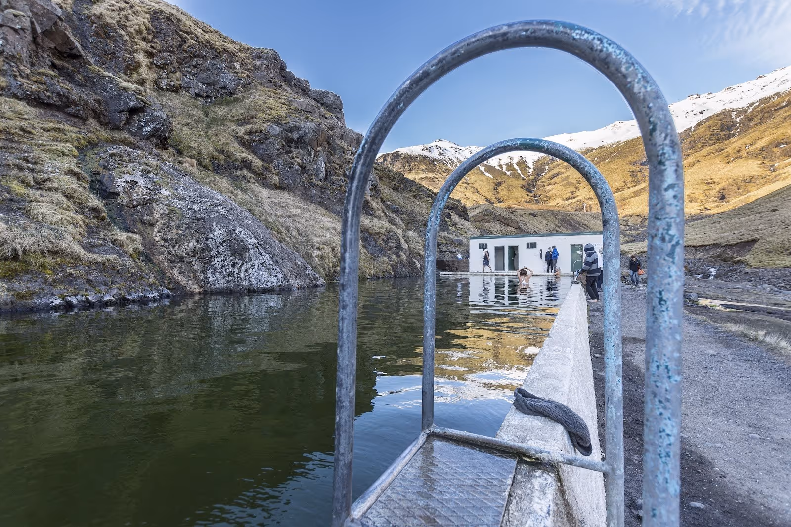 Close-up of the metal handrail at Seljavallalaug hot spring pool with a white building and rocky hills in Skogar Iceland.