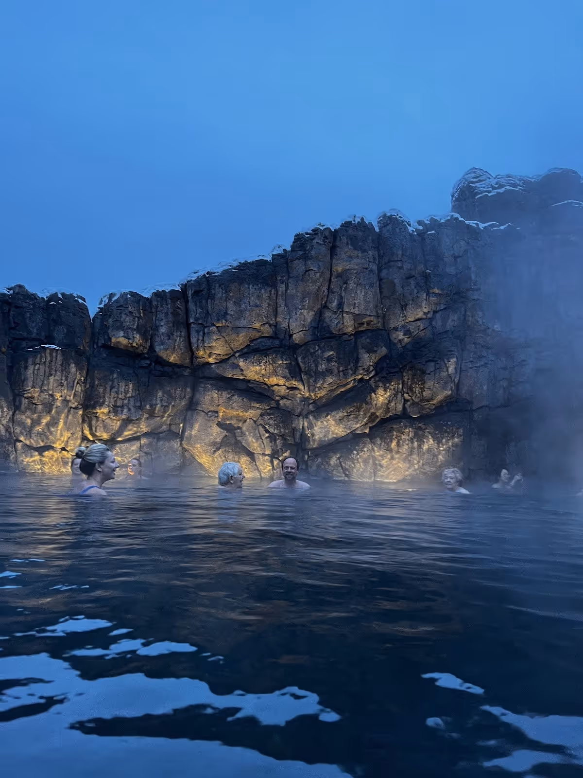 Several people relax in the mineral-rich hot spring pool with rocky cliffs in the background and gentle steam floating above the water at S.