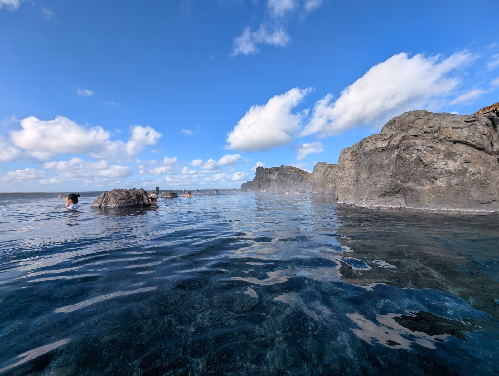 Visitors soak in the natural outdoor hot spring surrounded by large rocks with steam rising under a clear blue sky at Sky Lagoon near Reykj.