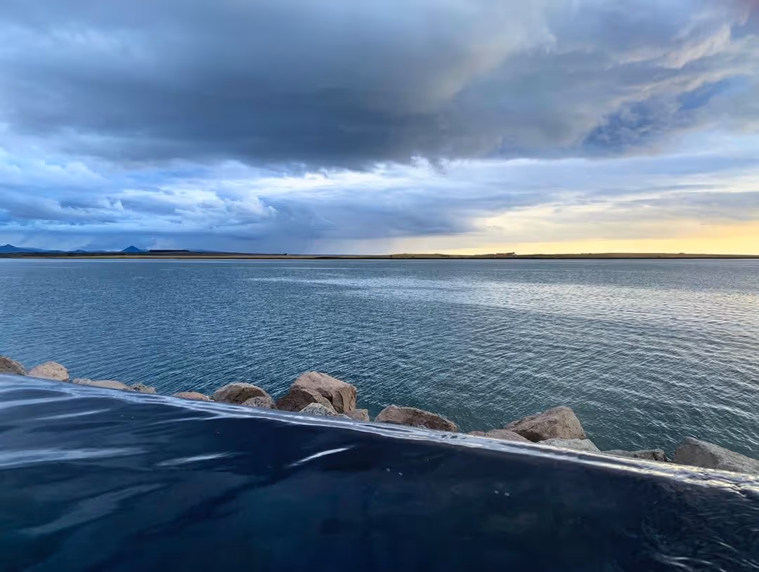 View of the dark water edge of the hot spring pool looking out across a calm large body of water with distant hills under moody clouds at S.