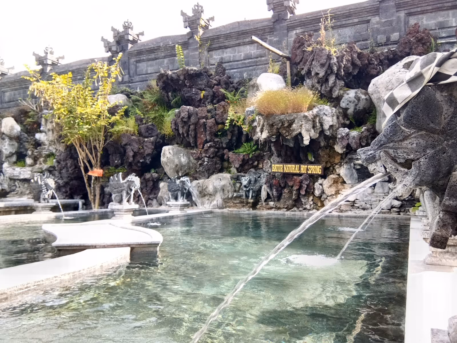 Hot spring pool with natural volcanic rocks and water spouting from stone sculptures at Batur Natural Hot Spring Bali.