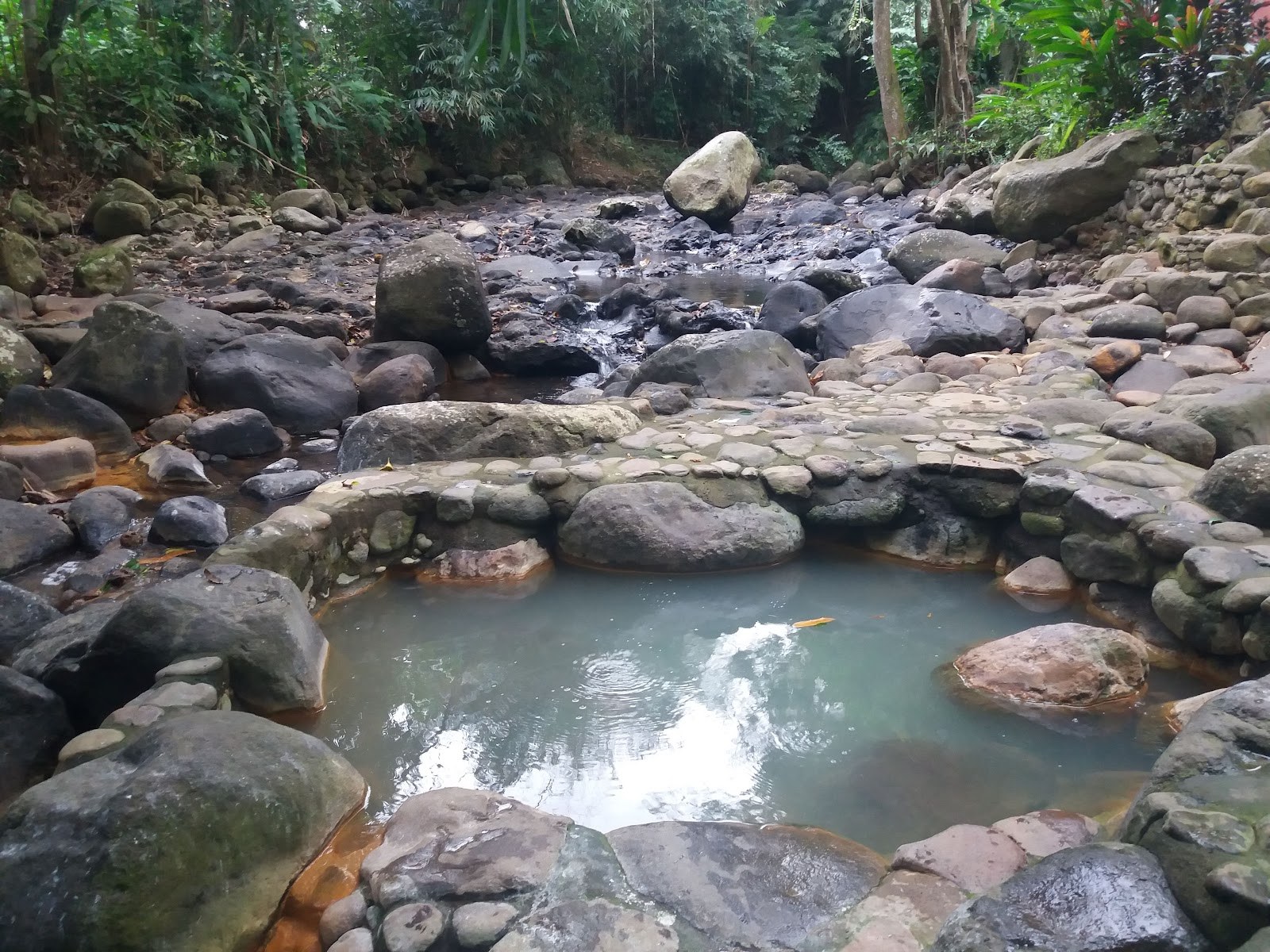 Natural hot spring pool enclosed by round stones along a rocky riverbed with dense forest trees near Tabanan Bali.