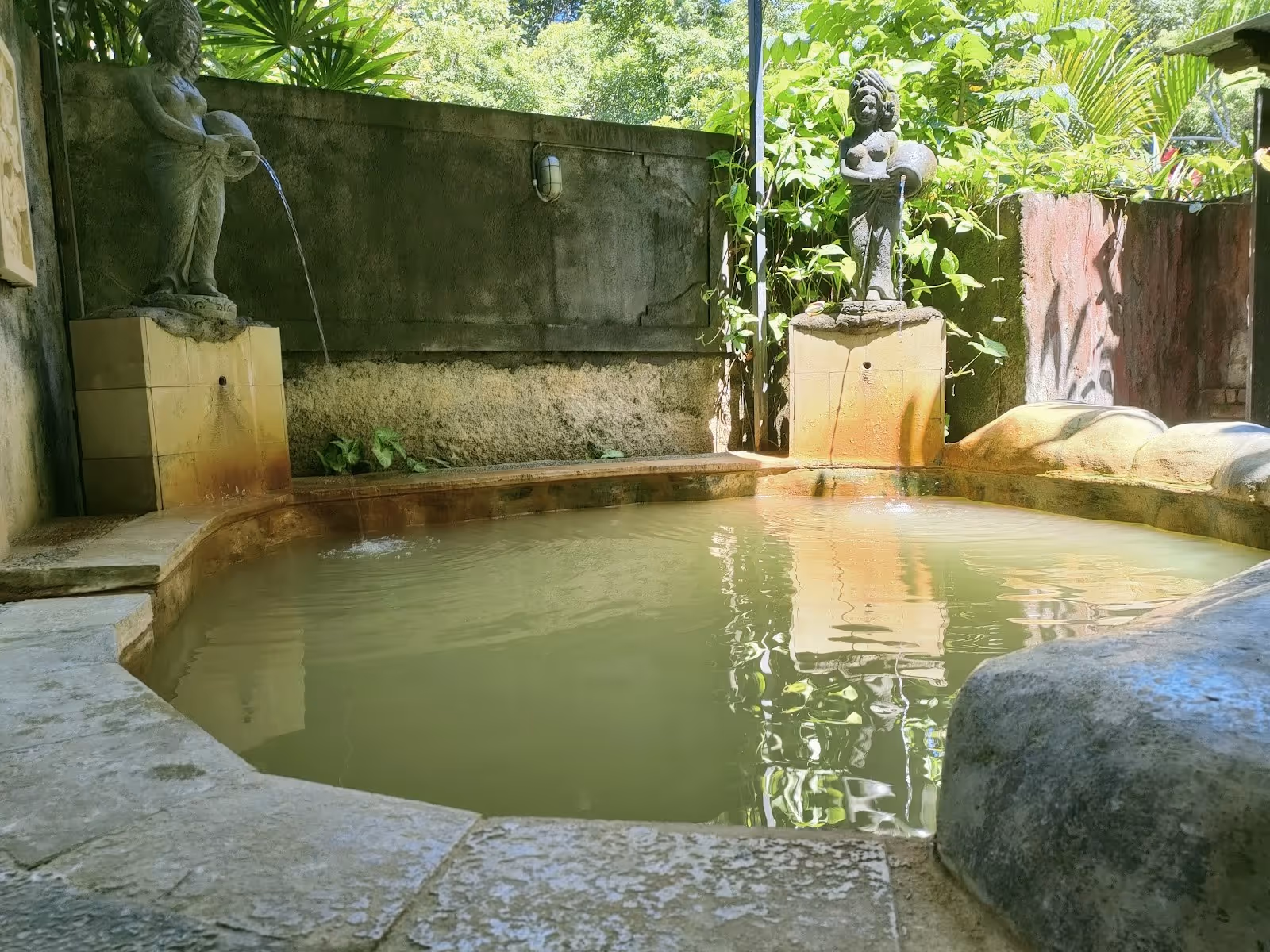 Stone-edge mineral hot spring pool with water fountains shaped like statues flowing gently among green tropical foliage.