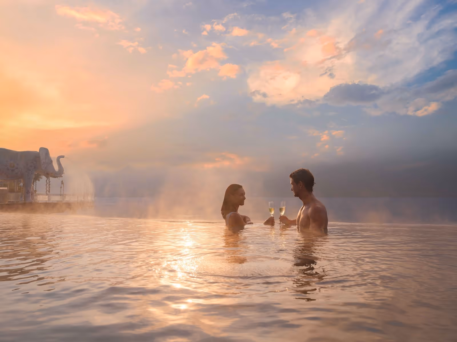A couple enjoying champagne in a steaming hot spring pool at sunset with a large elephant statue nearby in Kintamani Bali.