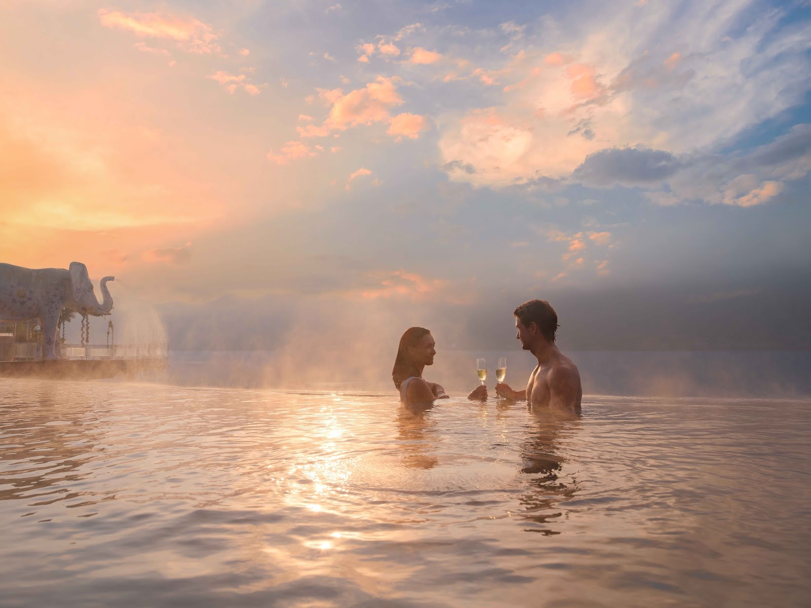 A couple enjoying champagne in a steaming hot spring pool at sunset with a large elephant statue nearby in Kintamani Bali.