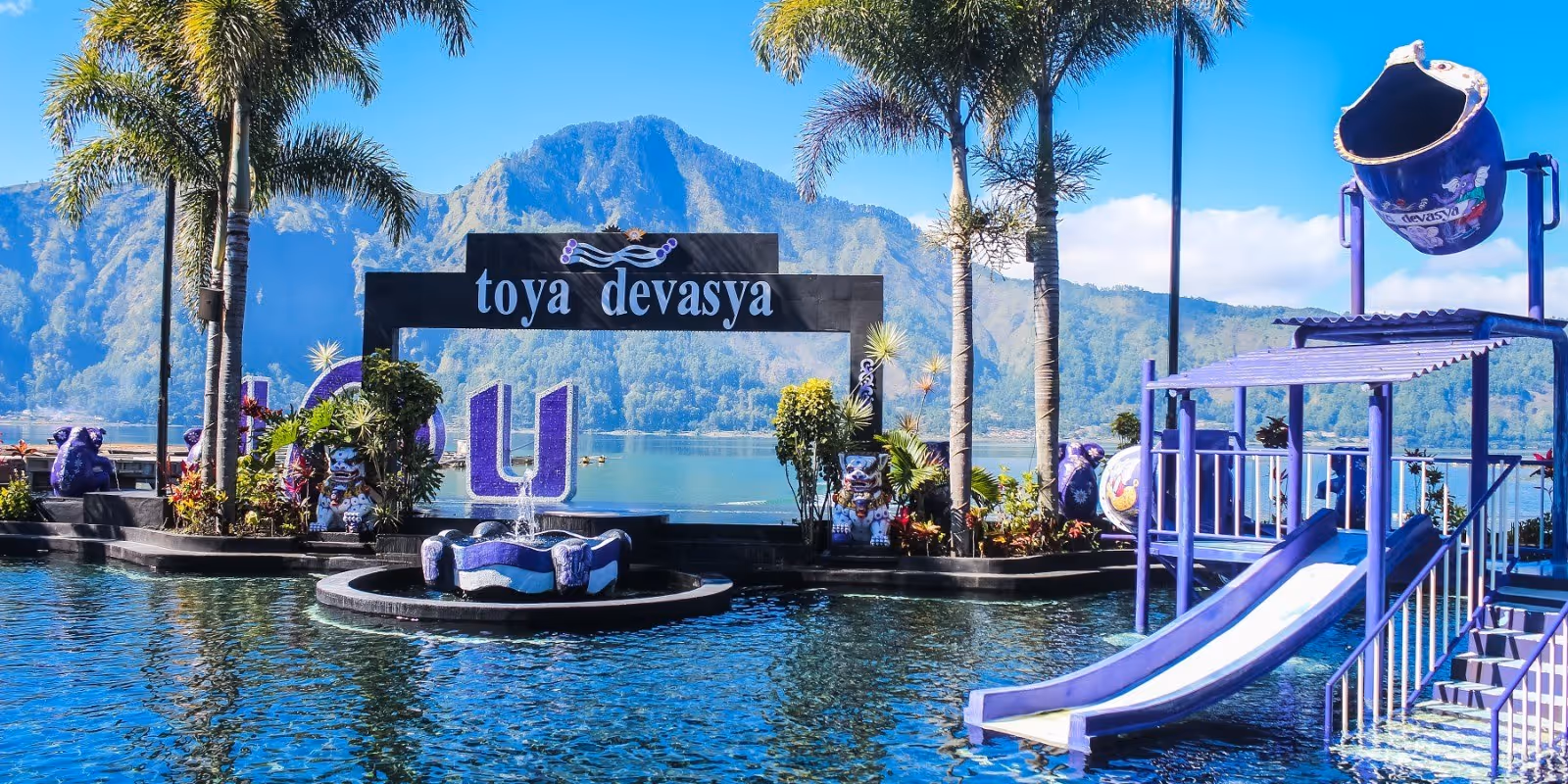 Outdoor mineral pool at Toya Devasya surrounded by palm trees and a sign with mountains visible in the background on a sunny day.