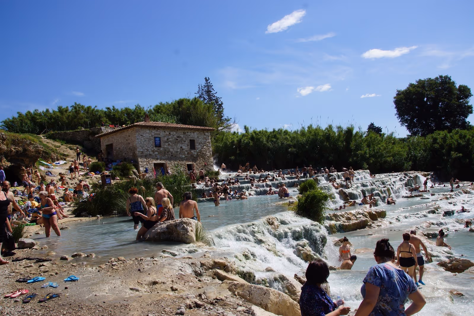 Many visitors enjoying the tiered hot spring soaking pools and stone building at Cascate del Mulino in Saturnia, Italy.