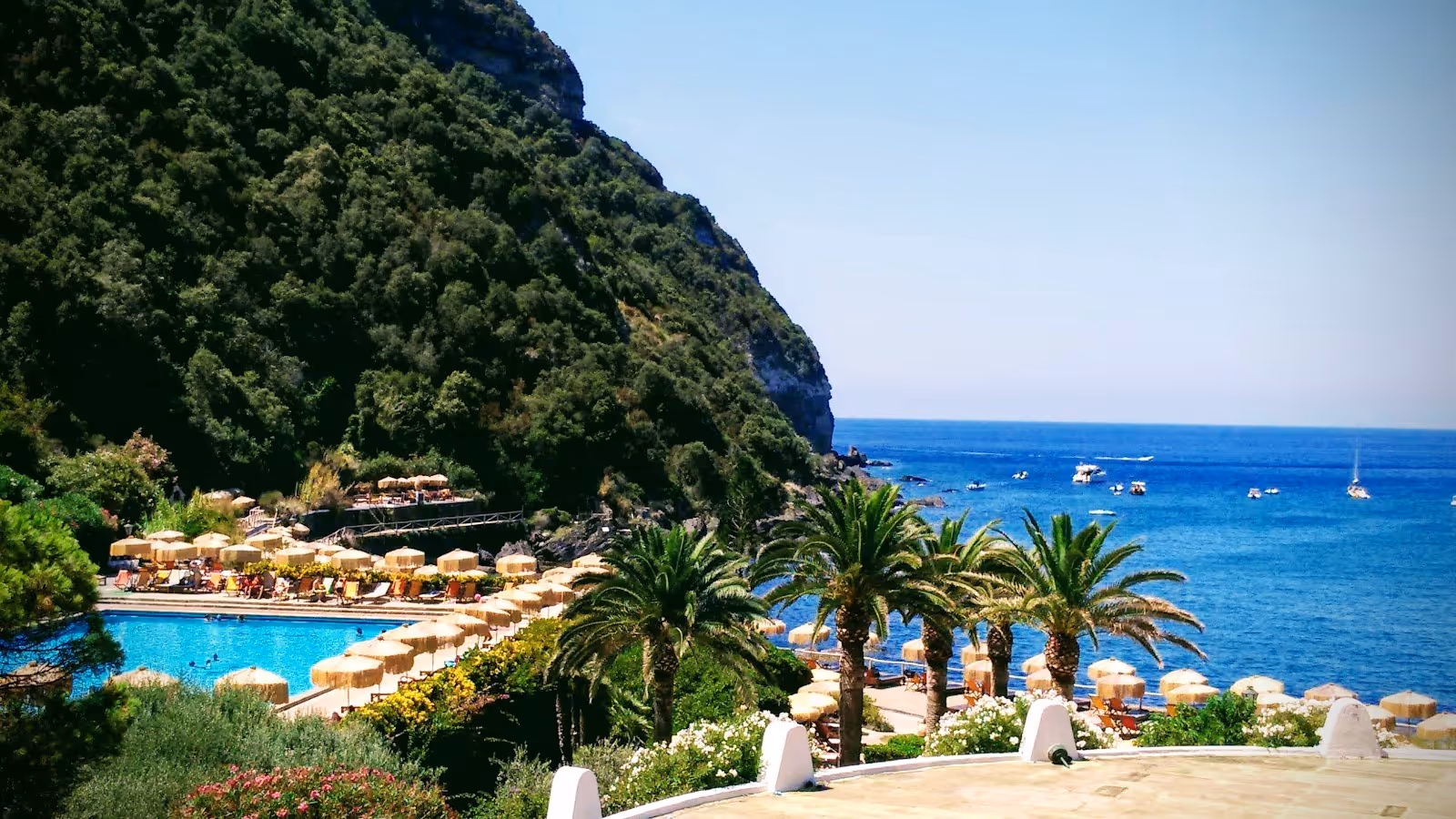 Giardini Poseidon Terme hot spring pool with palm trees and clear ocean water boats visible near Forio Ischia coastline in Italy.