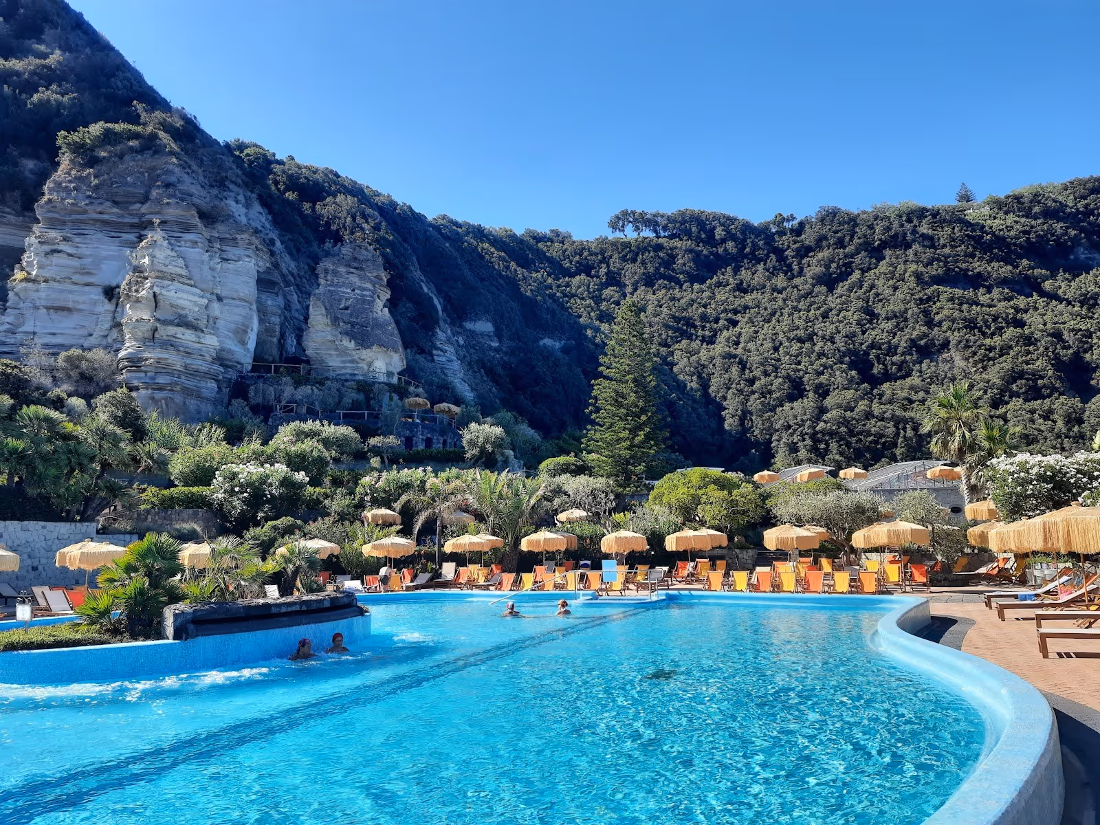 Curved outdoor mineral pool surrounded by lush green hills and yellow umbrellas at Giardini Poseidon Terme near Forio in Italy.