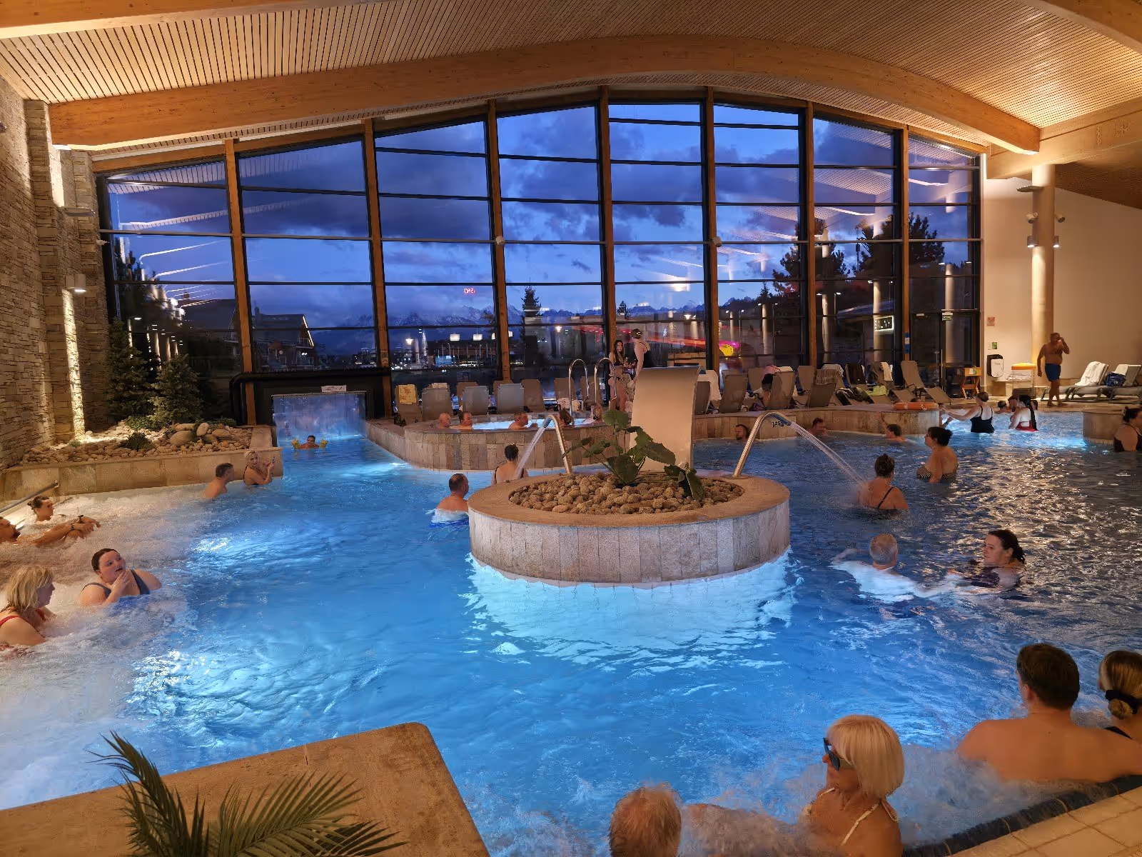 Indoor hot spring pool at Terma Bania Poland with many people relaxing near tall windows at dusk.