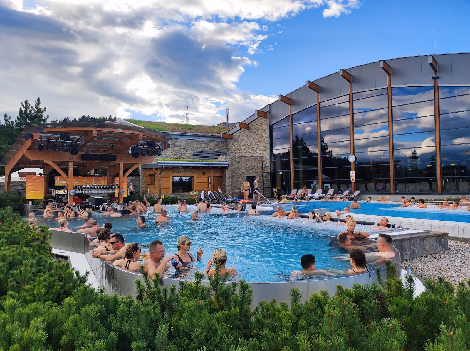 People sitting and enjoying water in a large outdoor hot spring pool with a building and green bushes in Poland.