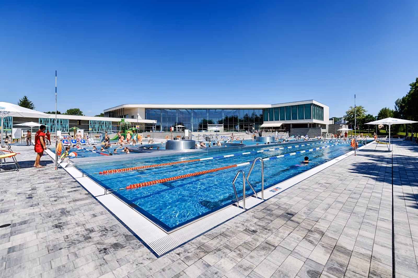 Large outdoor swimming pool with lanes and people relaxing near the glass-walled building of Termy Poddebice, Poland.