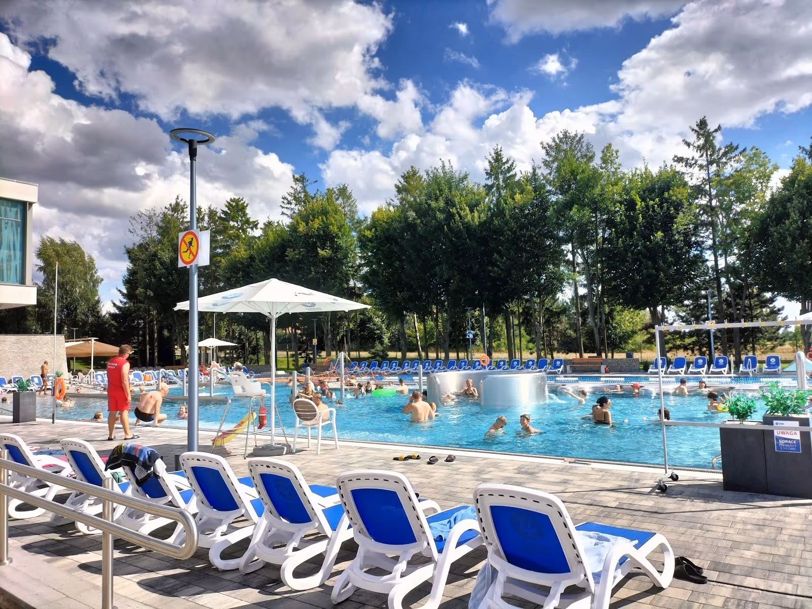 Crowded outdoor mineral pool features umbrellas and blue lounge chairs surrounded by trees at Termy Poddebice, Poland.