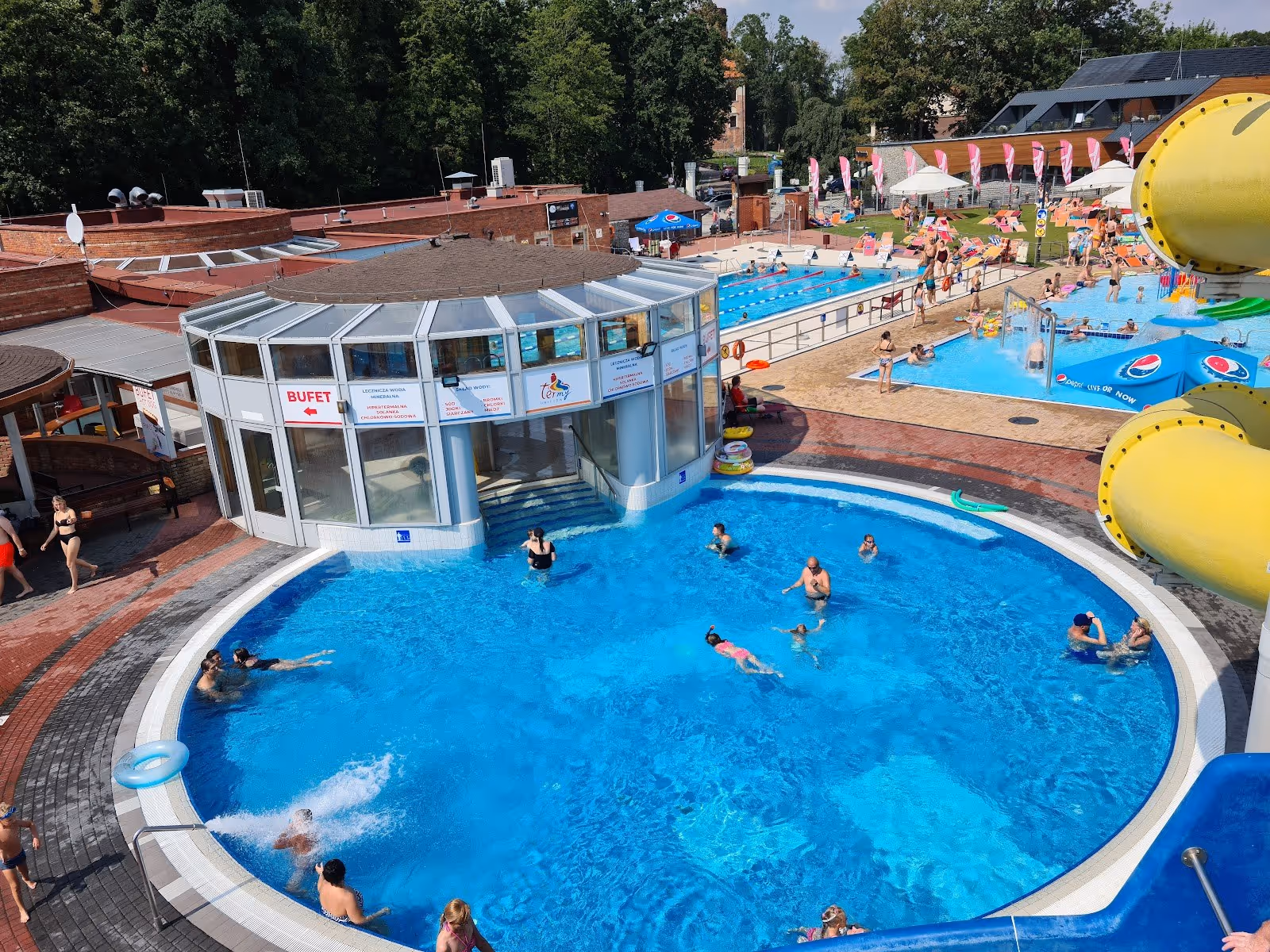Outdoor swimming pool at Termy Uniejow in Uniejow Poland with visitors enjoying clear blue water and a yellow water slide nearby.