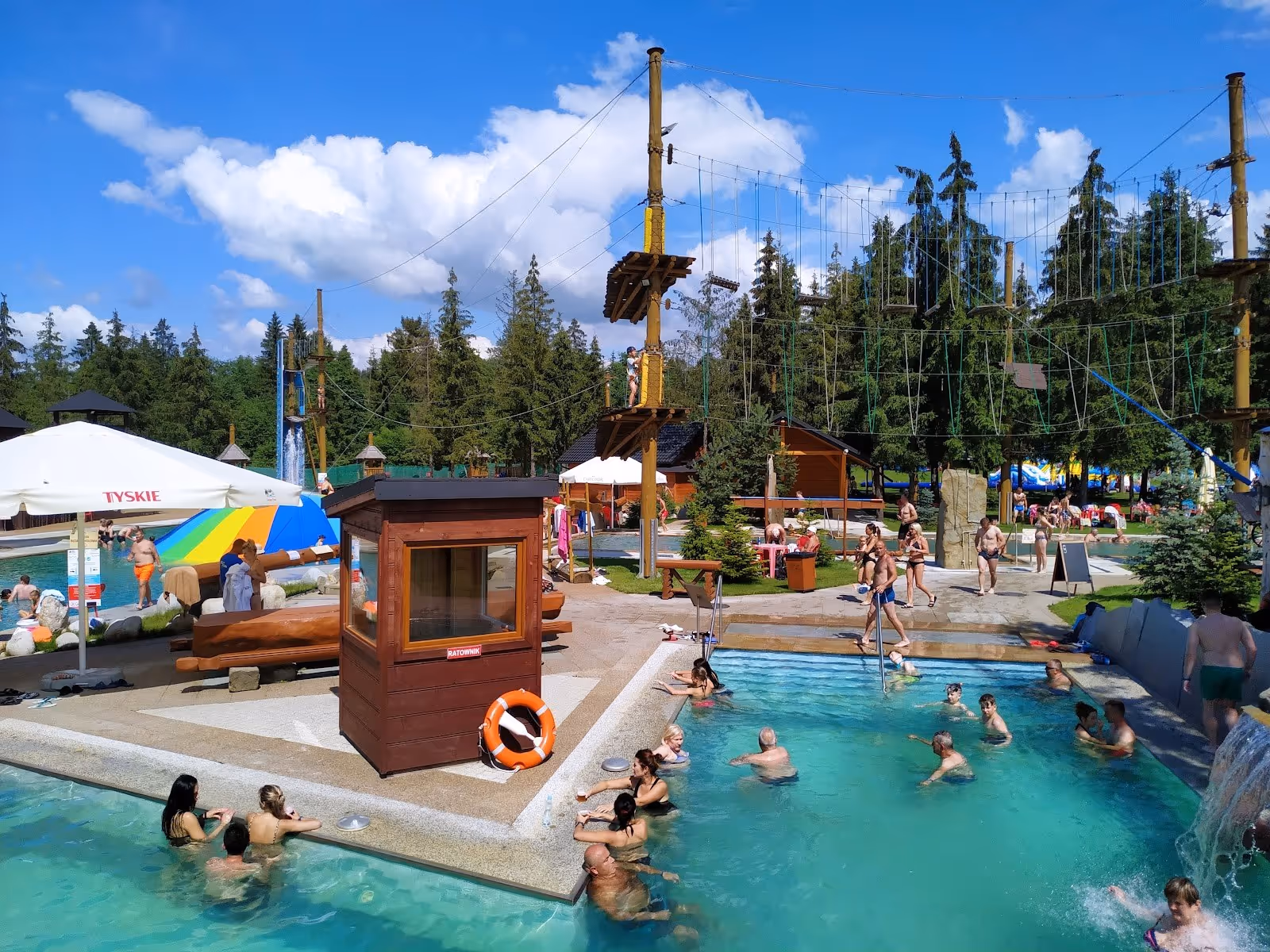 Group of people bathing in outdoor thermal pool with ropes adventure park visible in the background at Goracy Potok near Szaflary.