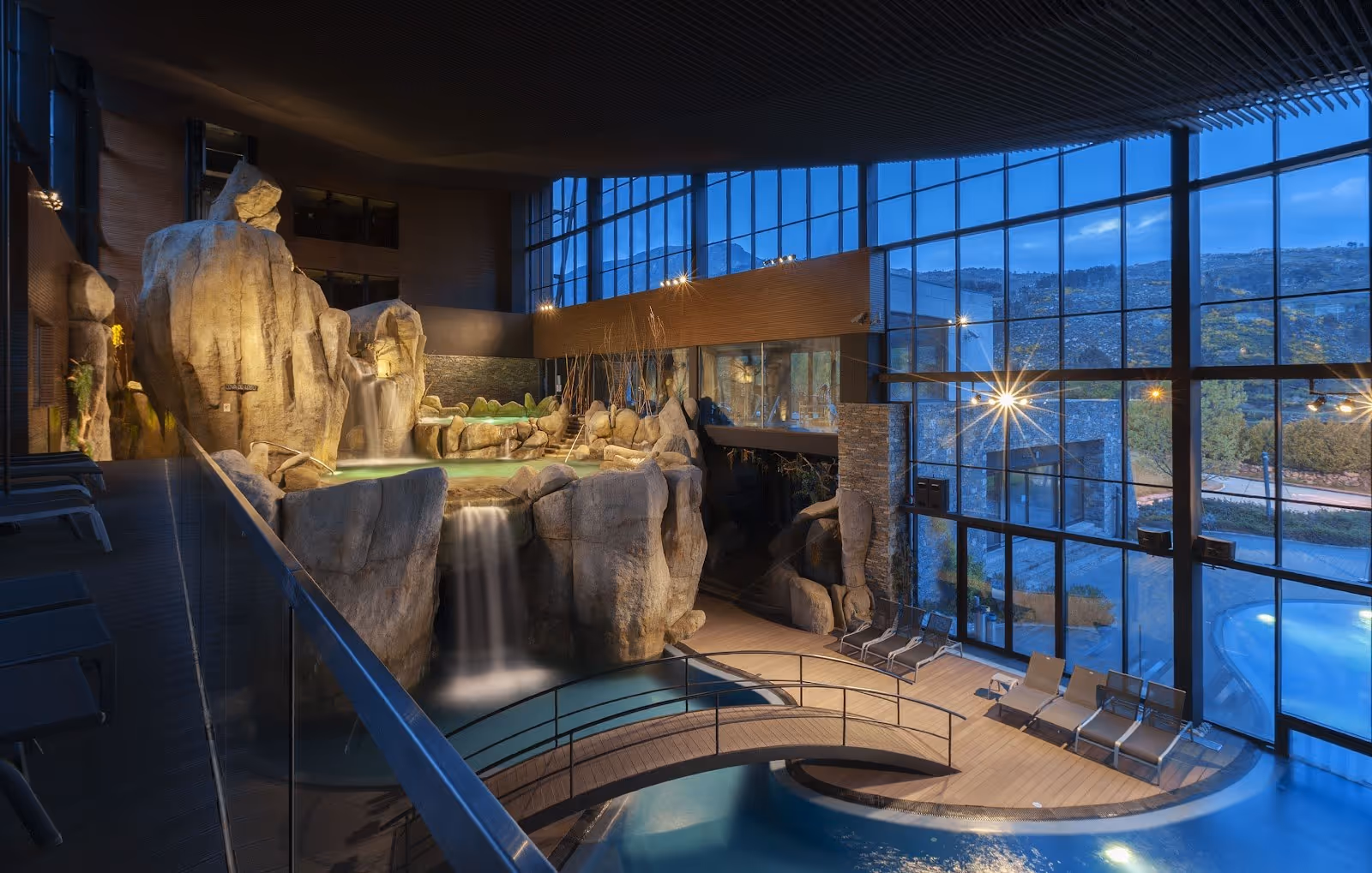 Indoor hot spring pool at Aquadome Termas de Unhais da Serra with rock waterfall and large glass windows showing a mountain view in portuga.