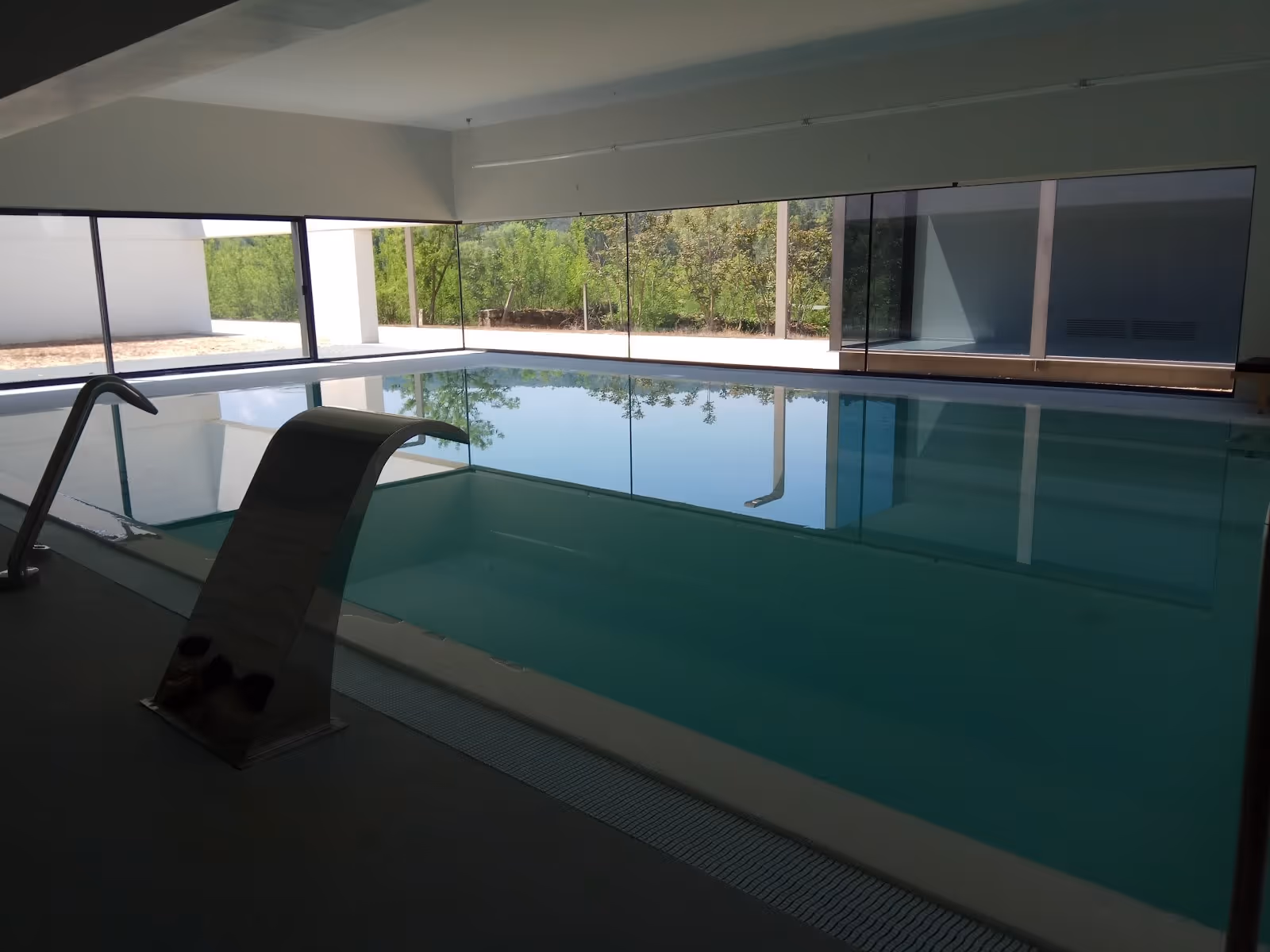 Calm indoor hot spring pool with a curved metal waterfall spout and a chair by the window at Balneario Pedagogico de Vidago in Portugal.