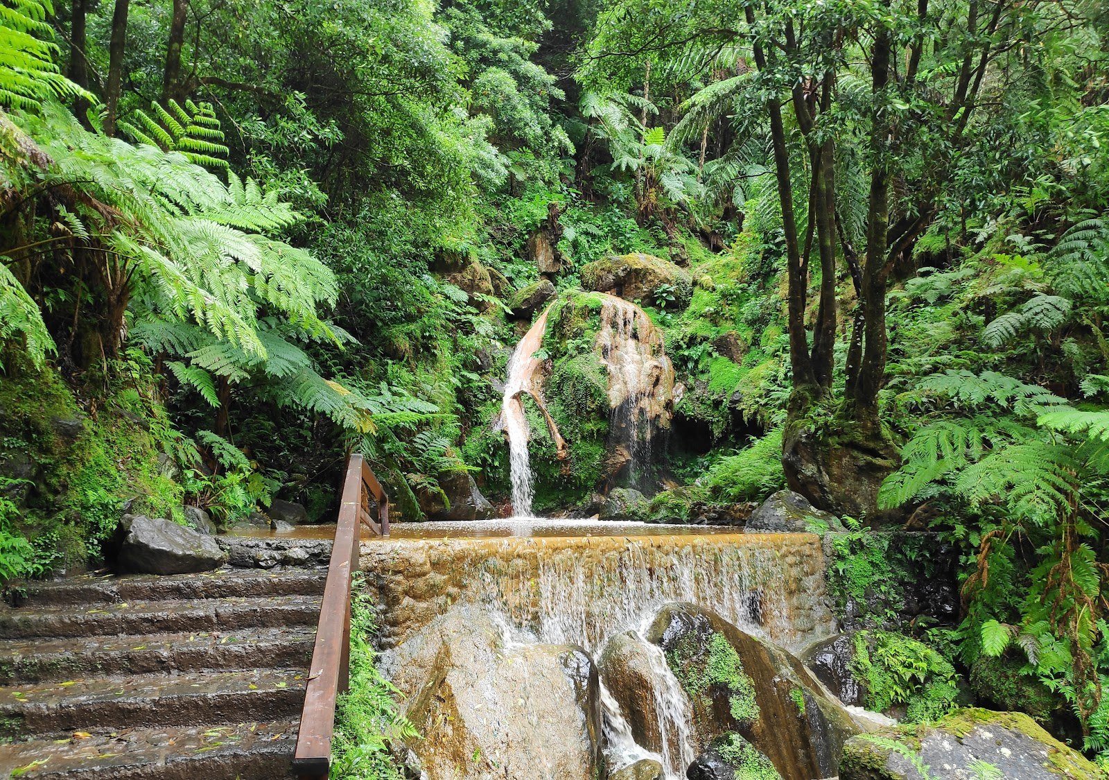 Caldeira Velha hot spring waterfall cascading over mossy rocks into a stone-edged pool with lush vegetation in Ribeira Grande Portugal.