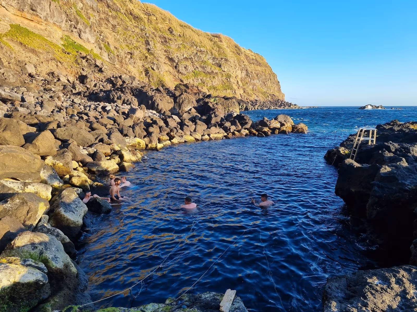 People soaking in a natural hot spring pool surrounded by volcanic rocks and cliffs at Ponta da Ferraria near Ponta Delgada Portugal.