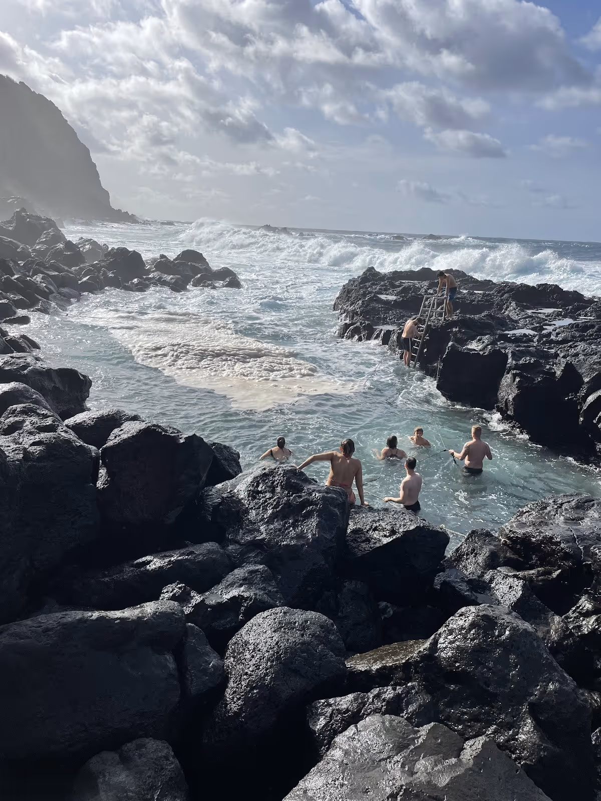 Visitors enjoy a calm natural pool with black volcanic rocks and ocean waves in the background at Ponta da Ferraria hot spring Portugal.