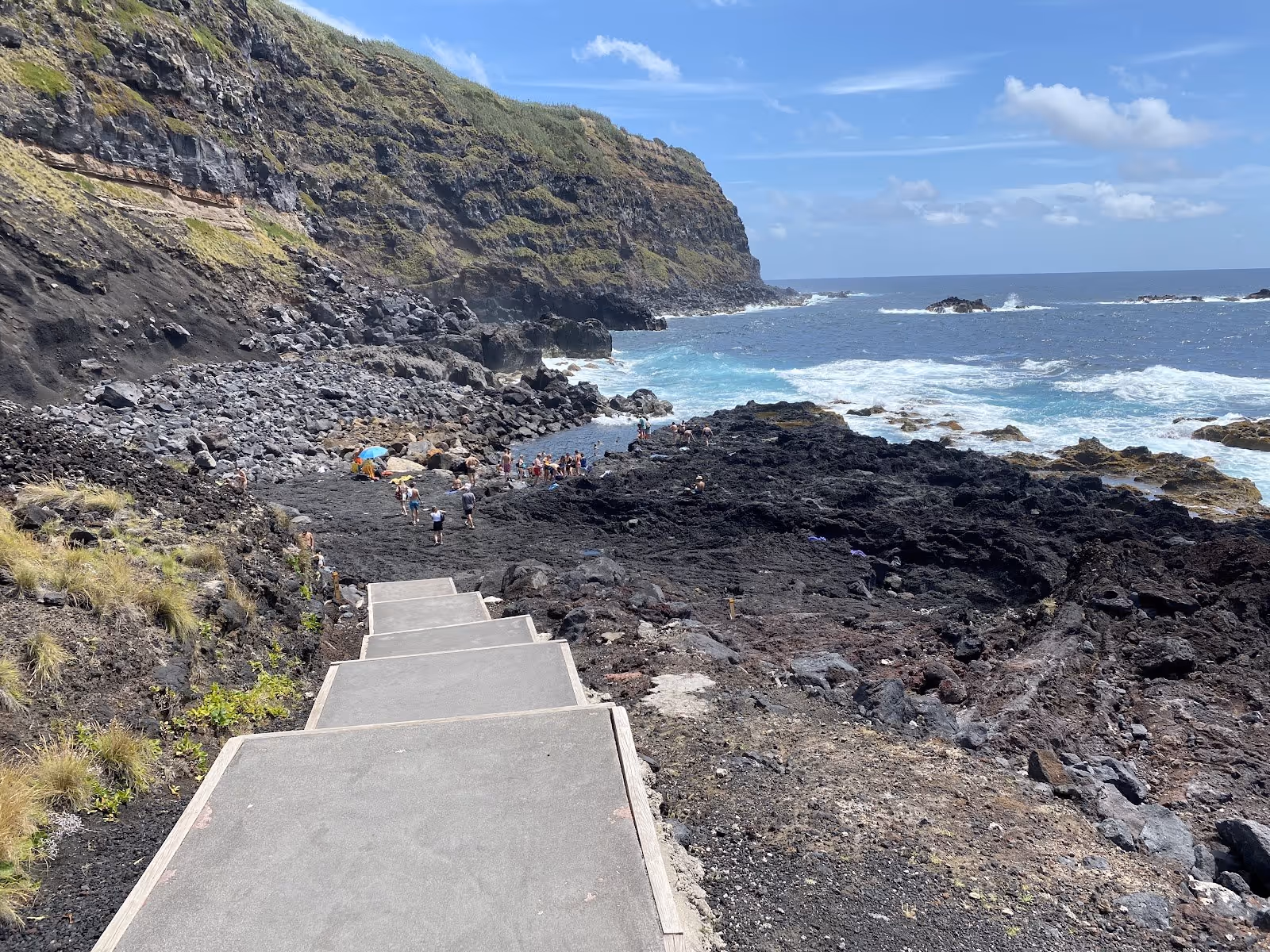 Group of people soaking in a rocky natural pool with waves crashing nearby under cloudy sky at Ponta da Ferraria near Ponta Delgada Portuga.