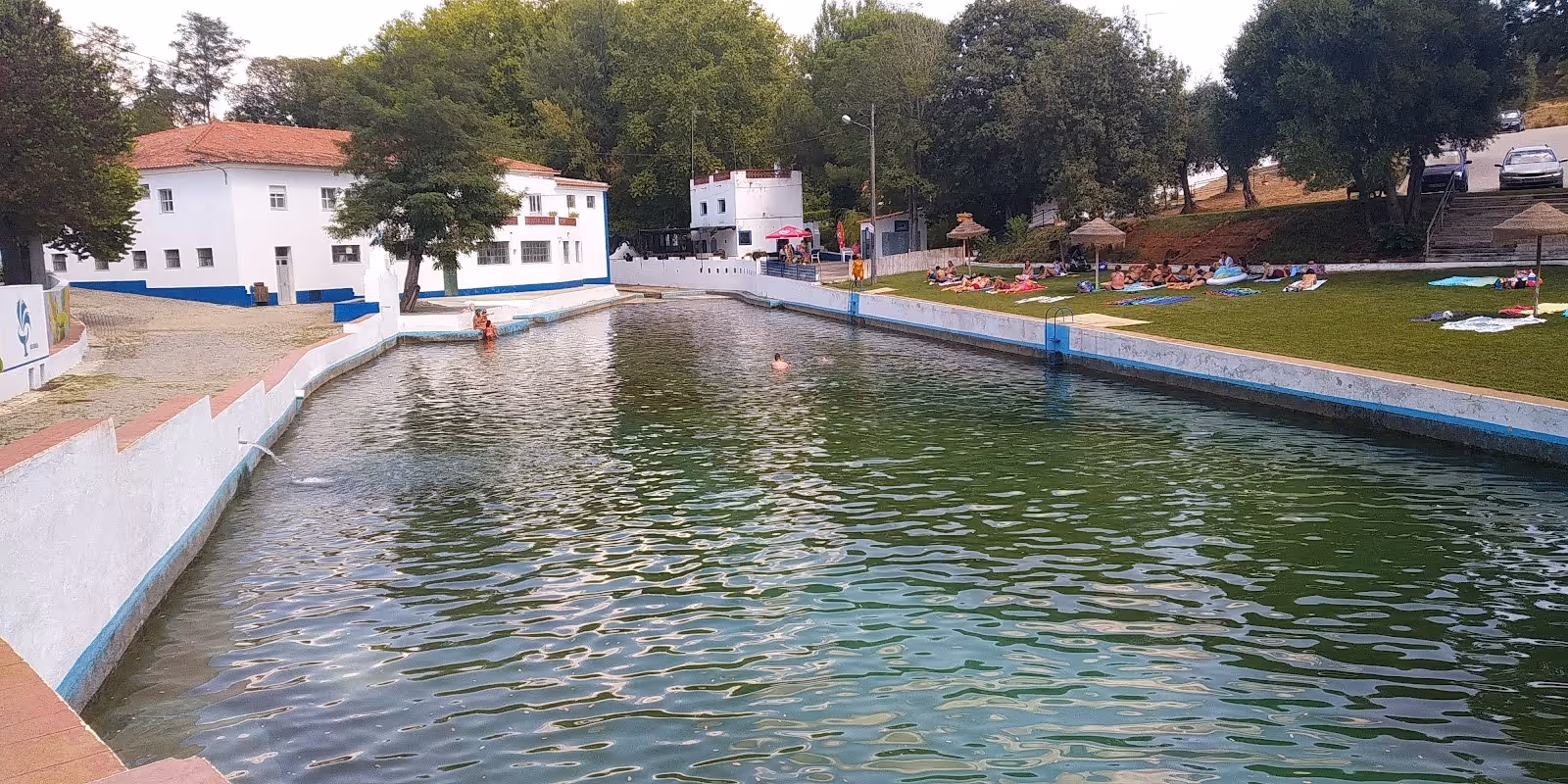 A long, rectangular outdoor hot spring pool with clear green water, surrounded by trees and white buildings at Termas da Sulfurea in Fronte.