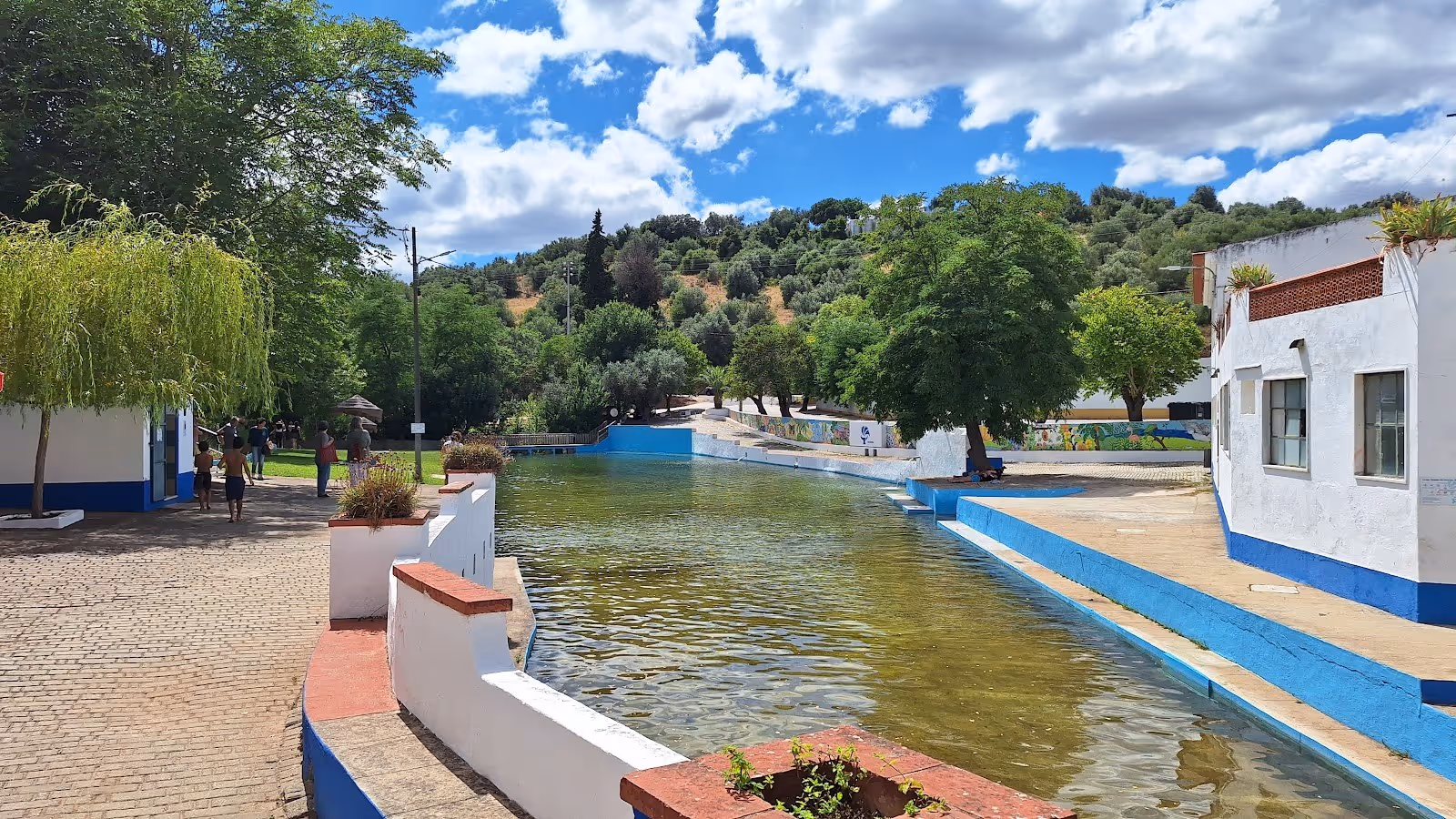 Outdoor soaking pool with a few people near white buildings painted with blue trim, set against a background of green hills at Termas da Su.