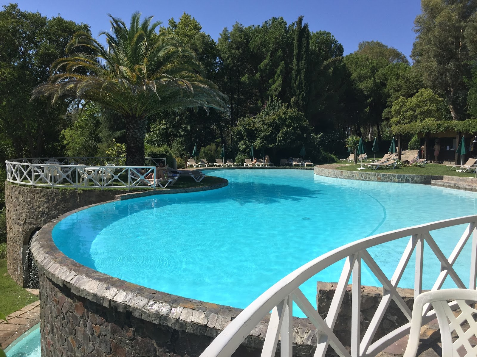Outdoor hot spring pool at Termas de Monfortinho with clear blue water surrounded by lush trees and scattered lounge chairs in Monfortinho.