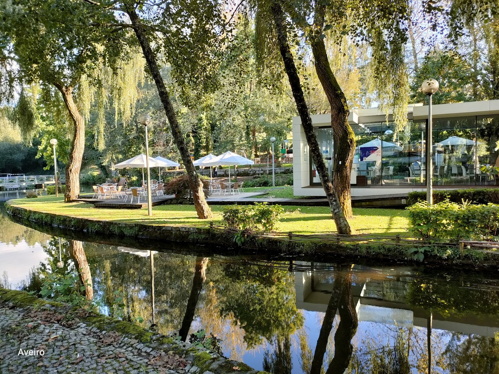 Green park area with white parasols and a glass cafe building beside a reflective water canal at Termas de S. Jorge by Santa Maria da Feira.