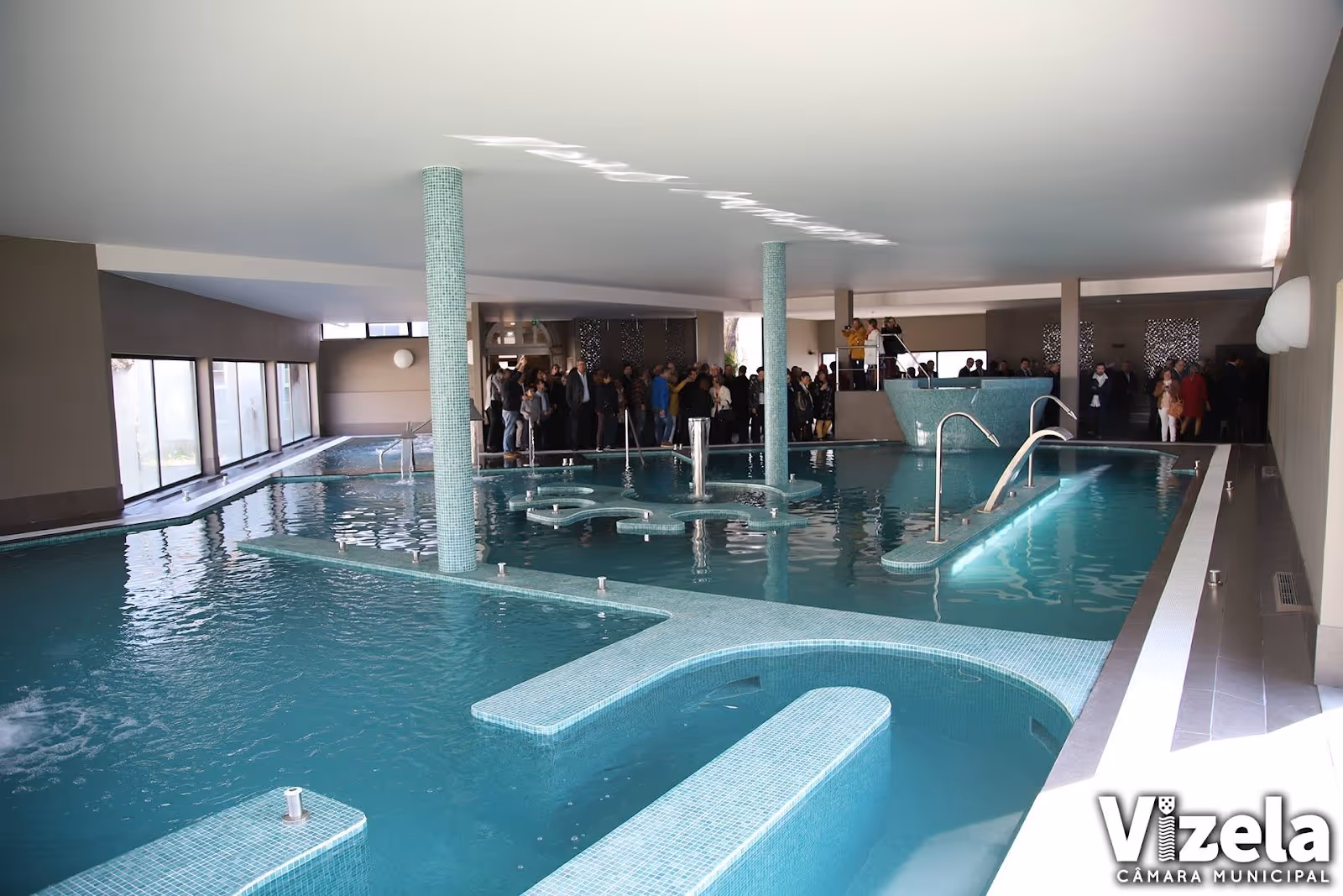 Indoor hot spring pool at Termas de Vizela in Portugal with multiple sections, smooth tiled edges, and gathered visitors in the background.