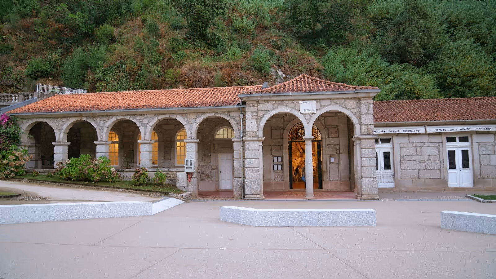 Stone spa building with multiple arches and a tiled roof surrounded by greenery at Termas do Geres in Geres Portugal.