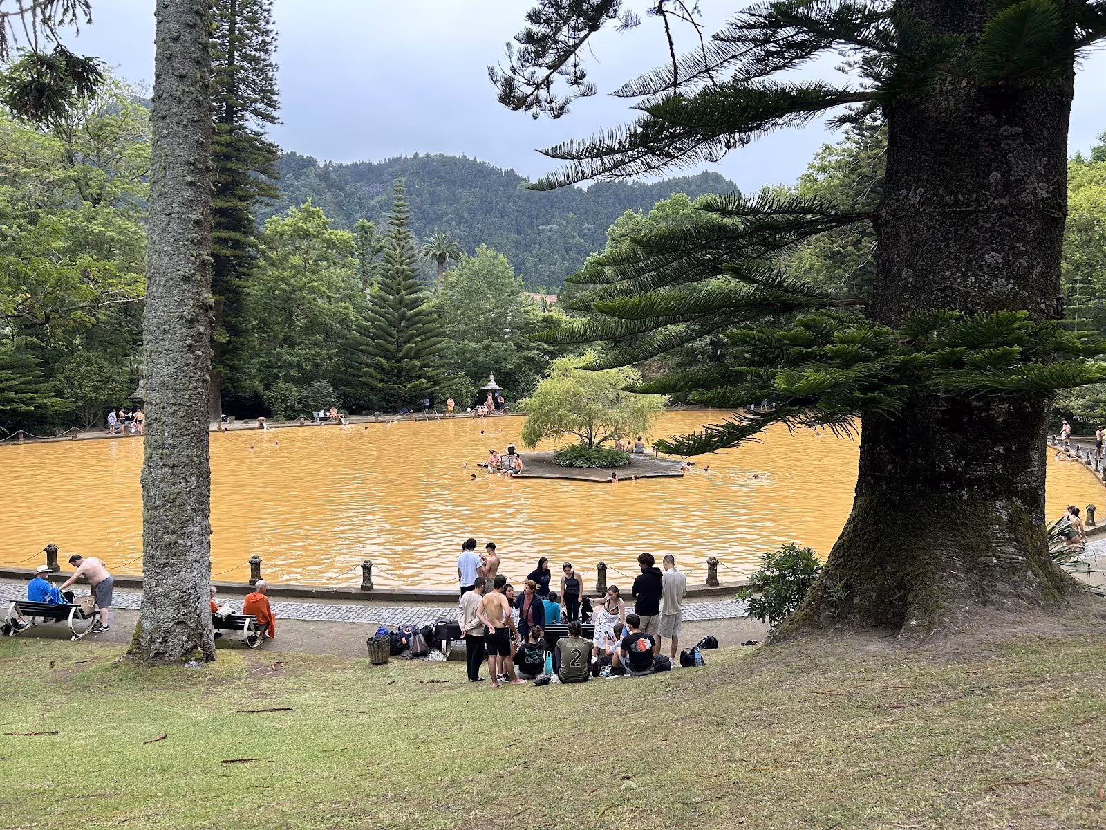 Large outdoor thermal pool with orange water and people sitting on grass near Furnas in Portugal at Terra Nostra Thermal Pool.