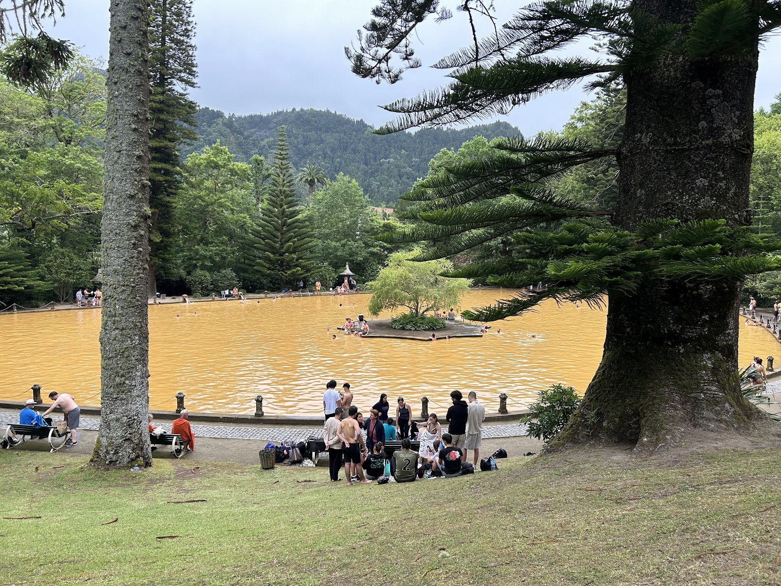 Large outdoor thermal pool with orange water and people sitting on grass near Furnas in Portugal at Terra Nostra Thermal Pool.