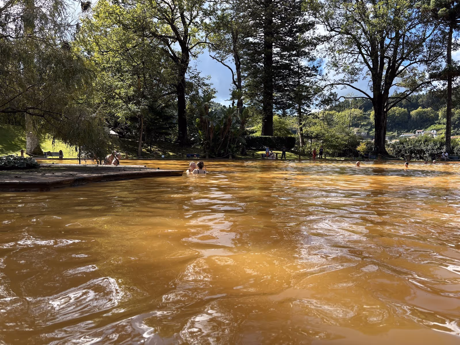 Orange mineral pool of Terra Nostra Thermal Pool with several people bathing, shaded by large leafy trees in Furnas, Portugal.