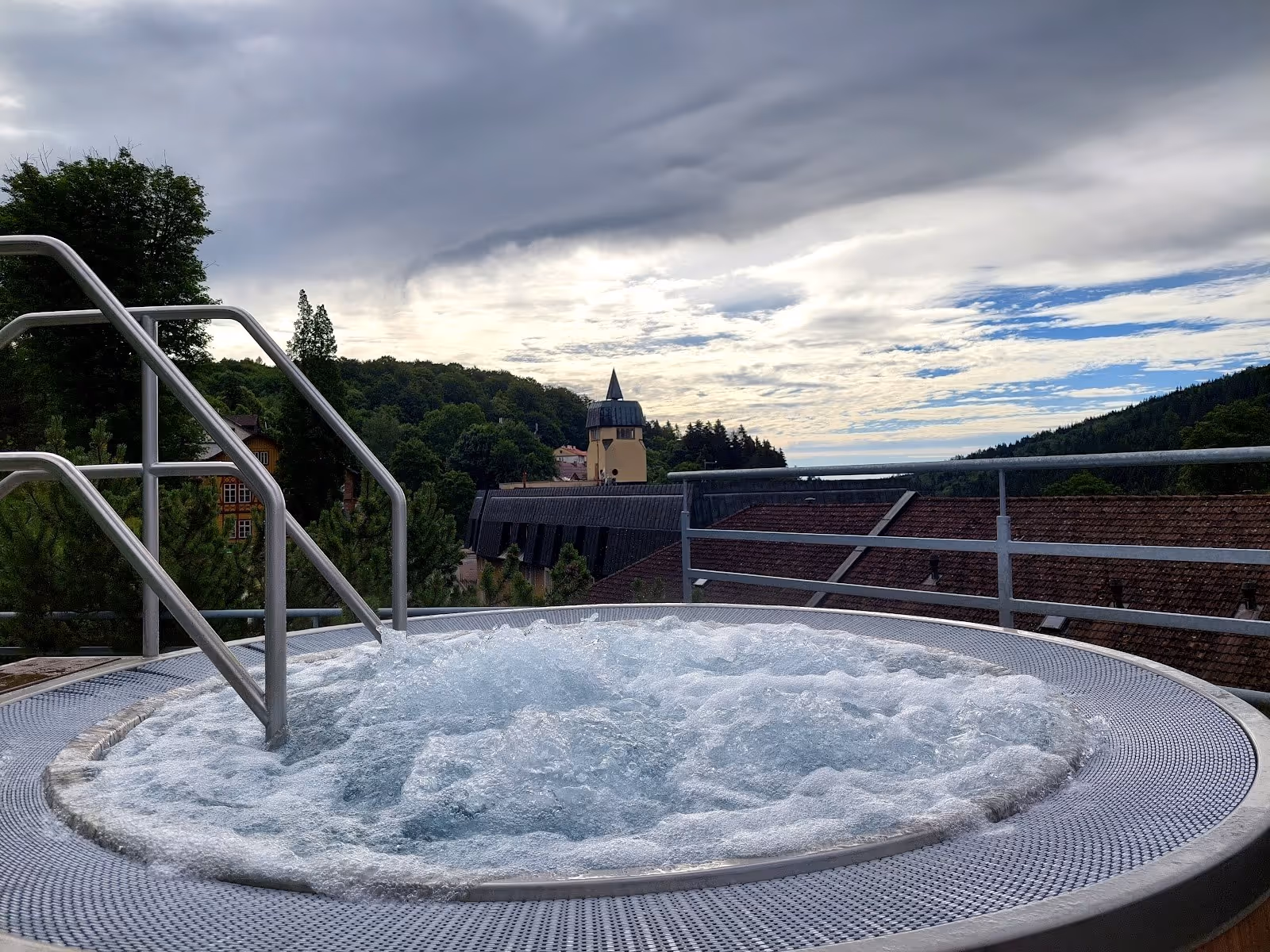 Outdoor bubbling hot tub with metal railing overlooking rooftops and a tower in Janske Lazne, czechia under cloudy sky.