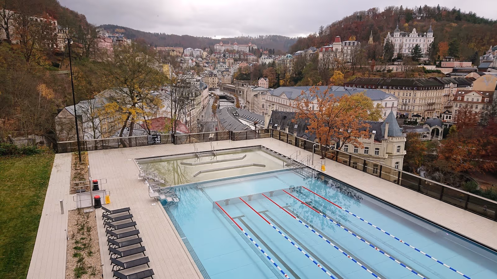 Two outdoor soaking pools with a fenced deck area and black lounge chairs overlooking the town of Karlovy Vary in Czechia during fall.