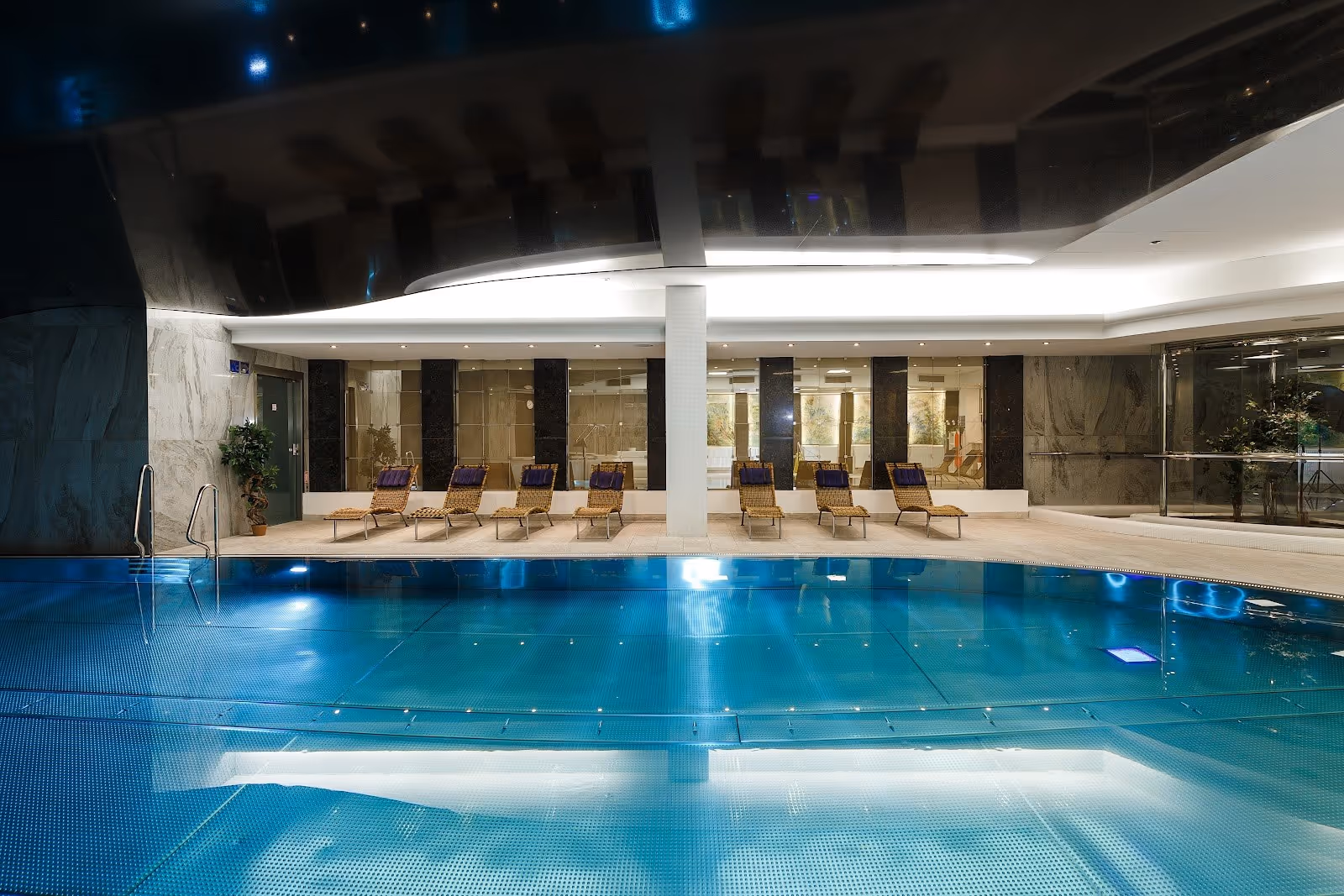 Indoor hot spring pool with lit blue tiles and woven lounge chairs aligned against a marble wall inside Hotel Thermal Pool in Karlovy Vary.